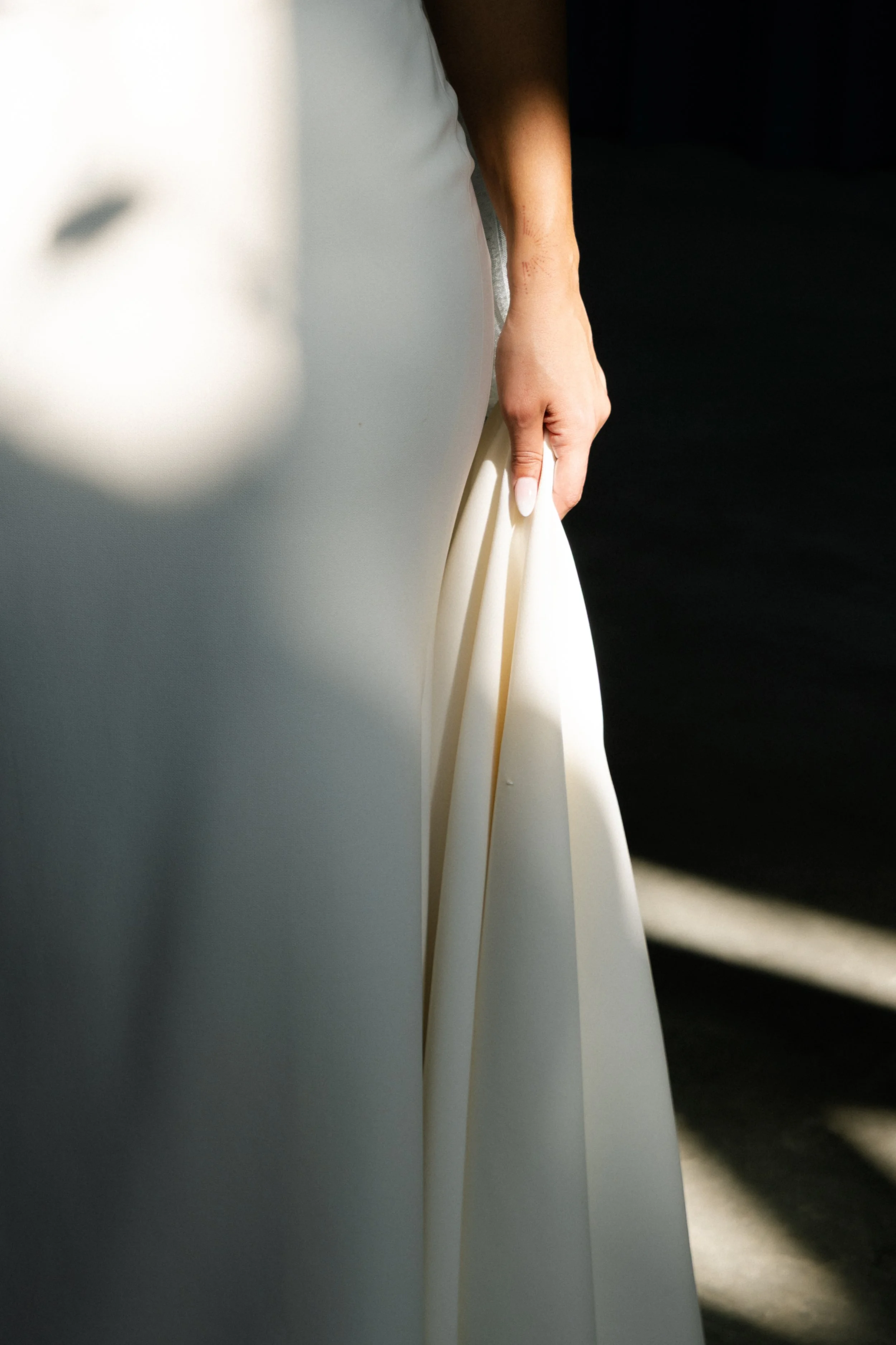 Close-up of a woman in a white dress holding the skirt, with soft sunlight casting shadows.