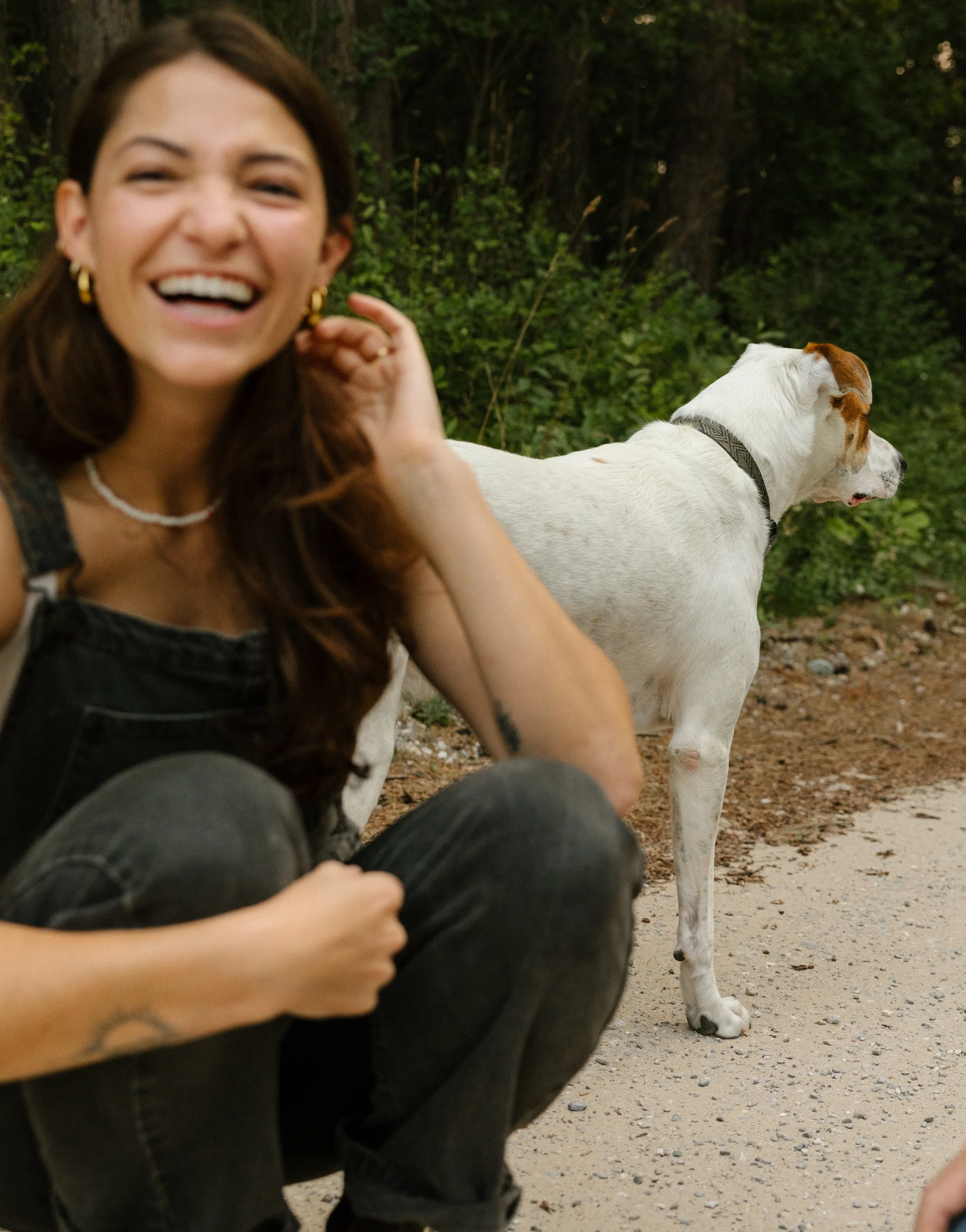 A woman laughing and sitting outdoors near a white dog with brown markings, in a wooded area.