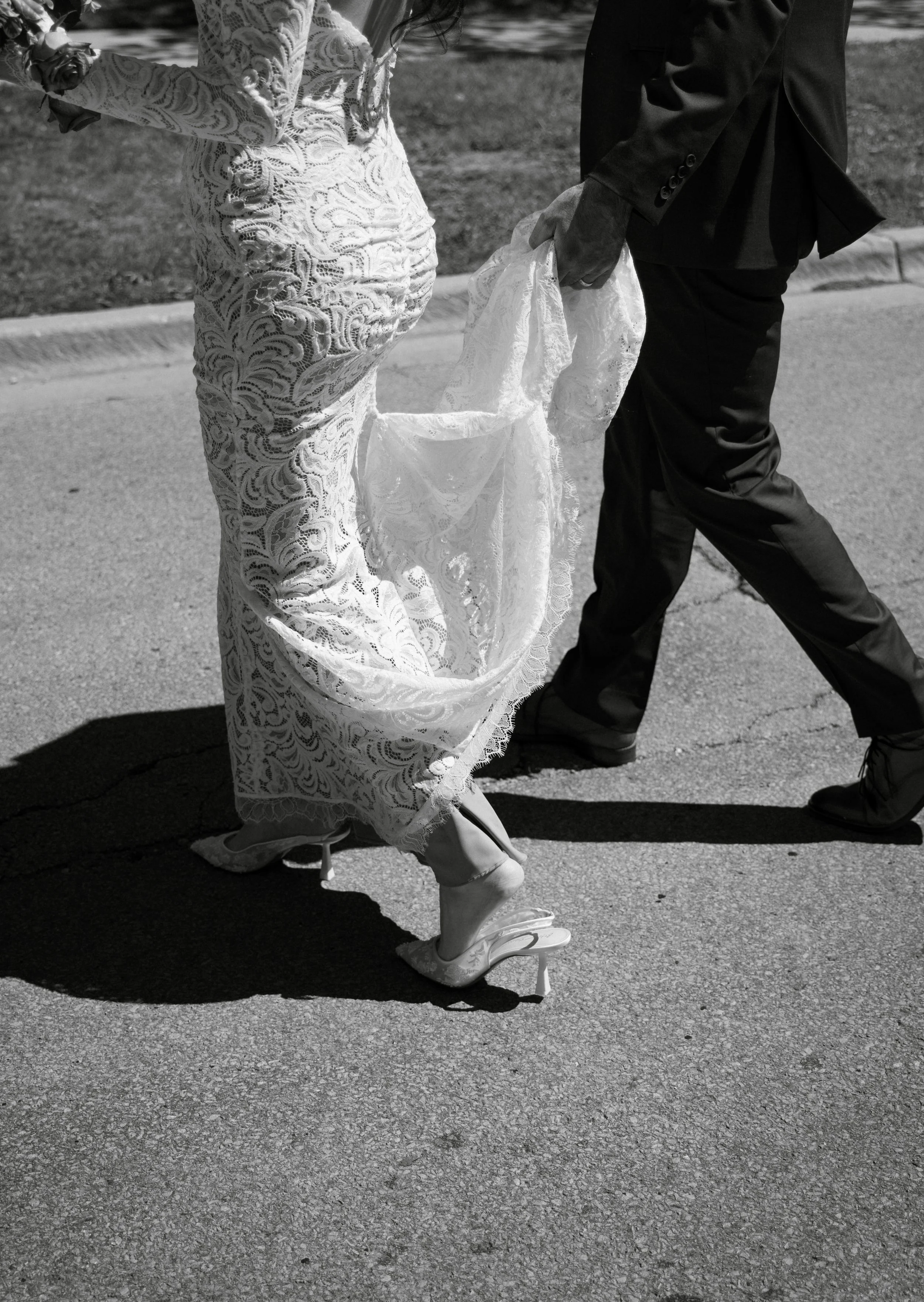 Black and white photo of a bride in a lace wedding dress and high heels, holding her gown with one hand, walking on a street with a groom in a suit, who is lifting part of her gown, on a sunny day.
