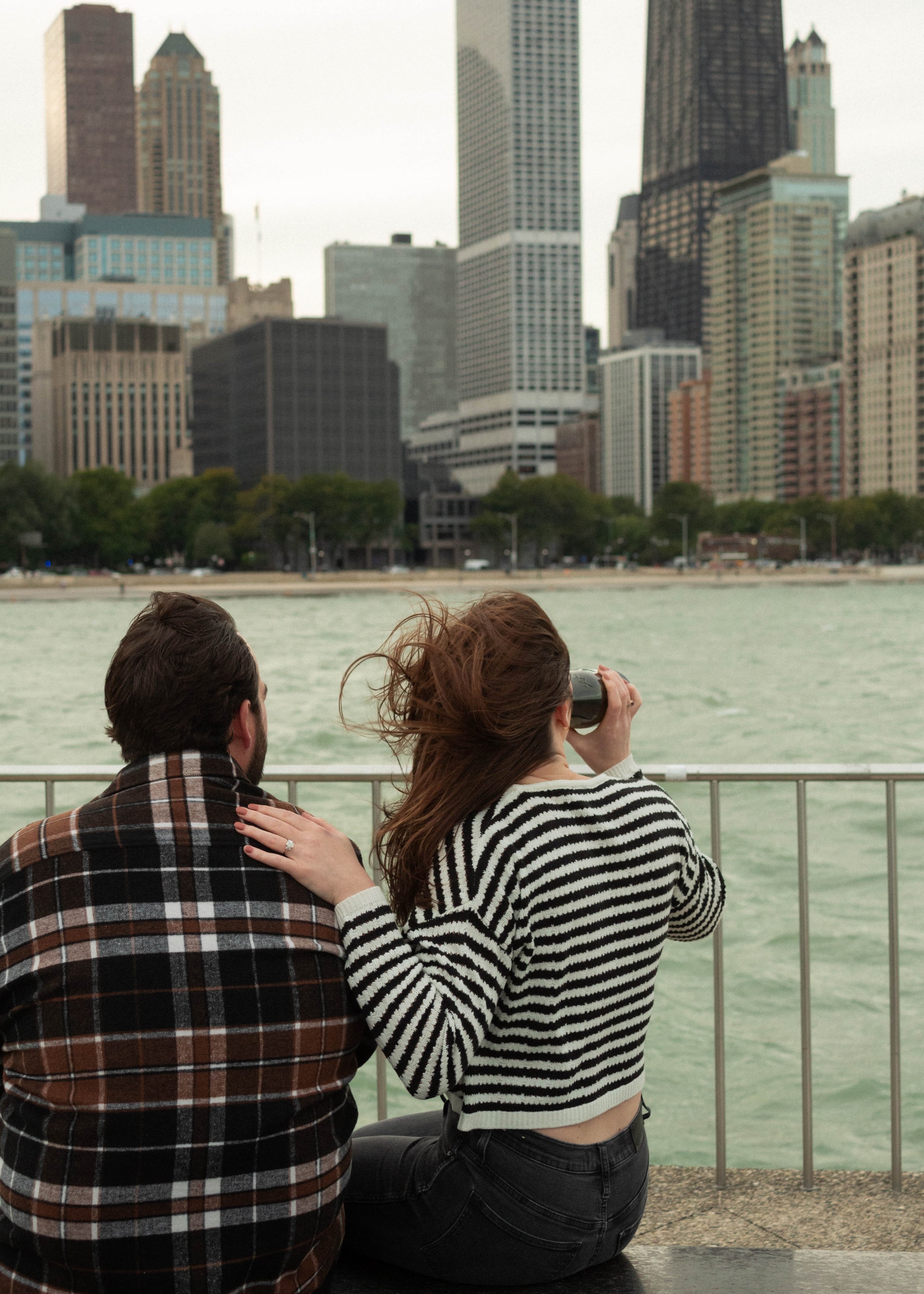 A woman with long brown hair wearing a black and white striped sweater is taking a photo with a camera, while a man in a plaid shirt sits beside her, on a waterfront with a city skyline in the background.