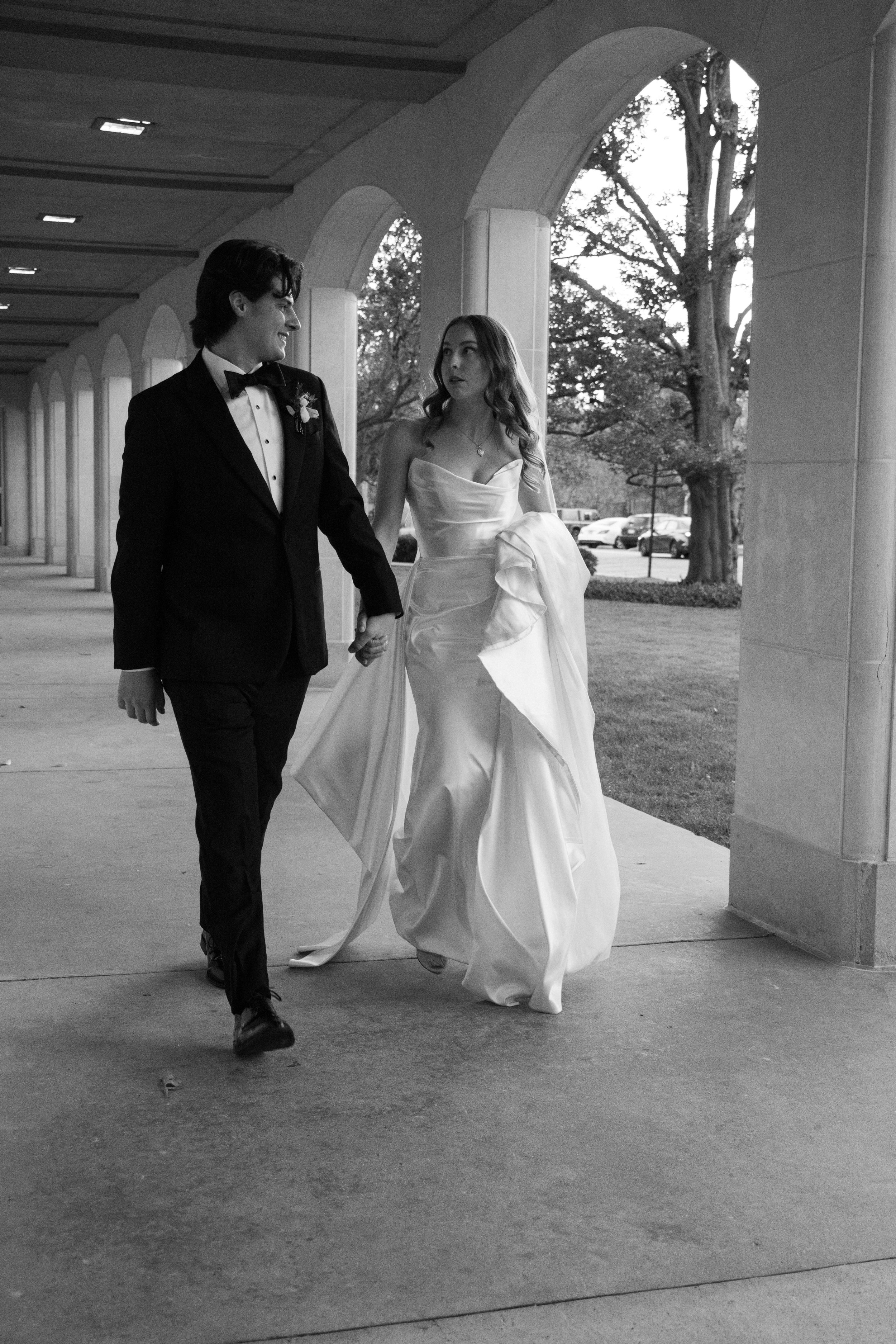 A bride and groom holding hands and walking under an arched walkway, with trees and parked cars in the background, in black and white.