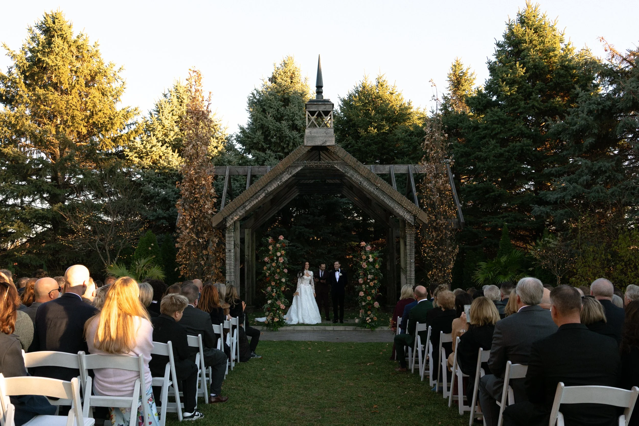 A wedding ceremony outdoors with guests seated on white chairs, a bride and groom standing under a wooden arch decorated with flowers, surrounded by tall trees.