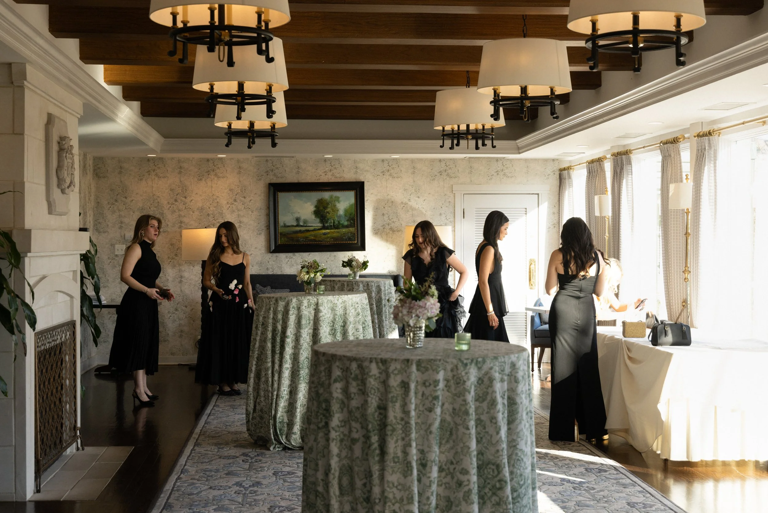 A quiet moment of a group of bridesmaids in elegant black dresses standing and sitting in a well-lit room with curtains, chandeliers, and a painting on the wall before the wedding ceremony