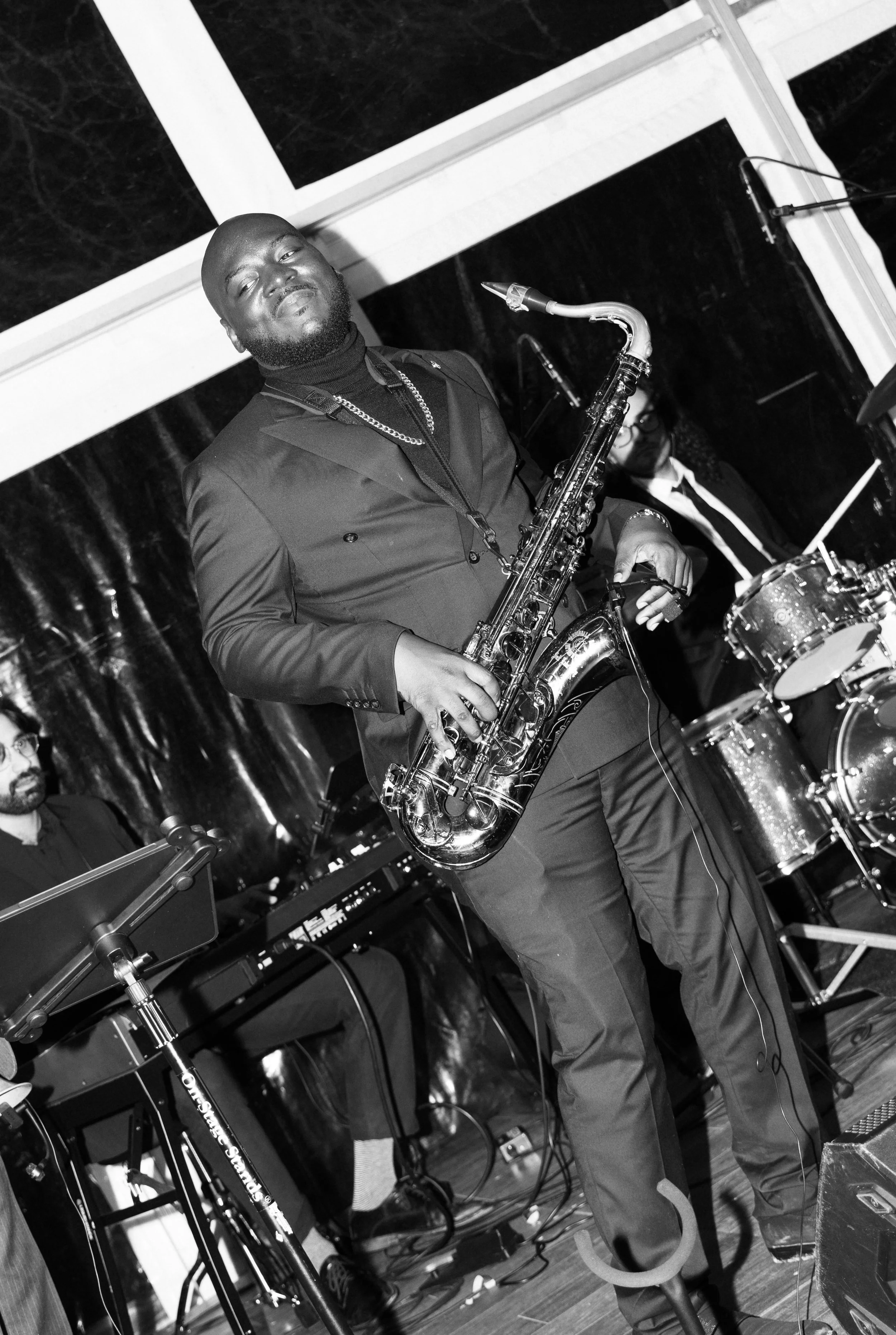 A man with a beard and stylish clothing playing a saxophone on stage, with a drummer and keyboard player in the background, during a live performance.