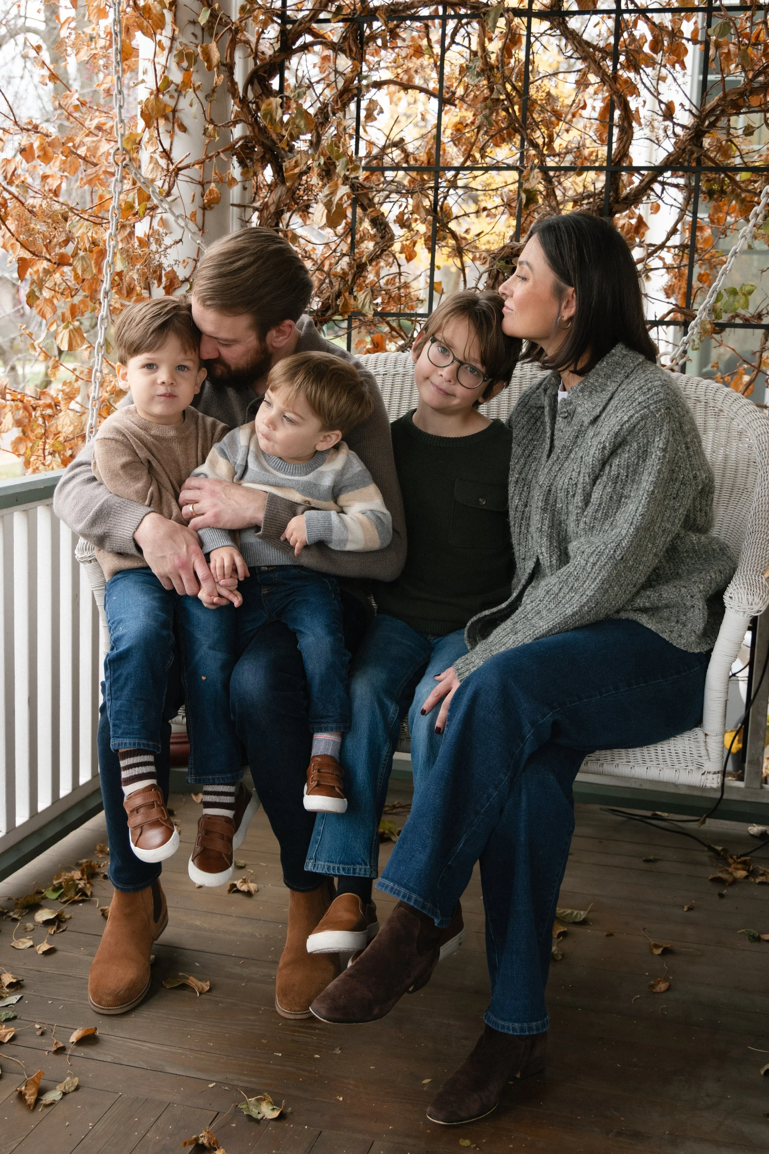 A family of six sitting on a porch swing surrounded by autumn leaves, with a man, a woman, and four young children, enjoying a fall day.