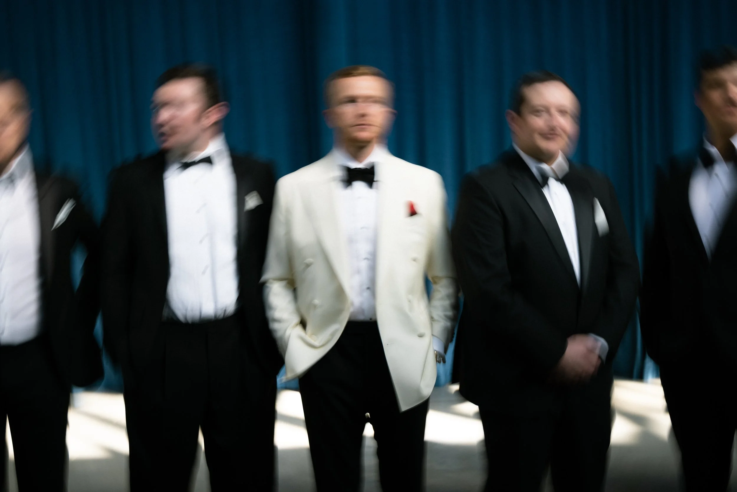 A group of men in black tuxedos and a man in a white tuxedo jacket standing in front of a blue curtain.