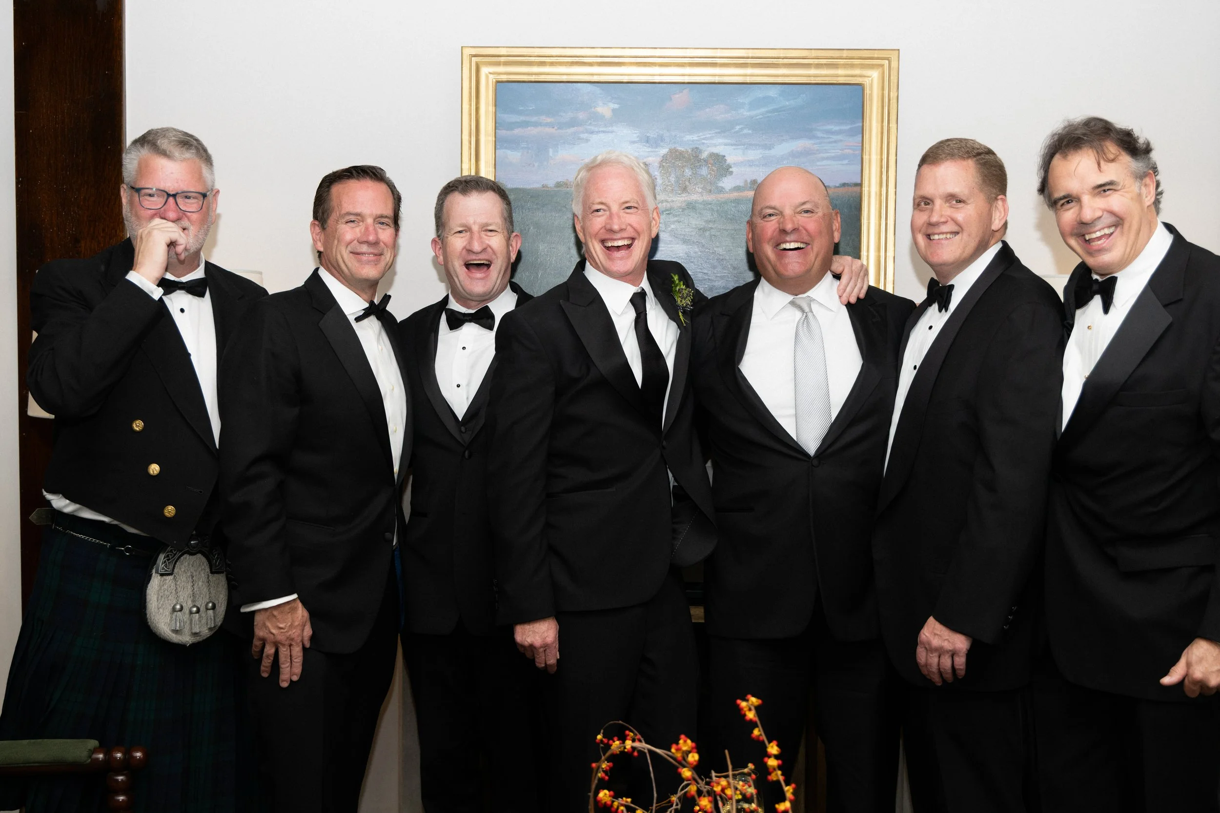 A group of seven men dressed in tuxedos, smiling and posing together at a formal event, standing in front of a framed landscape painting in a room with white walls.