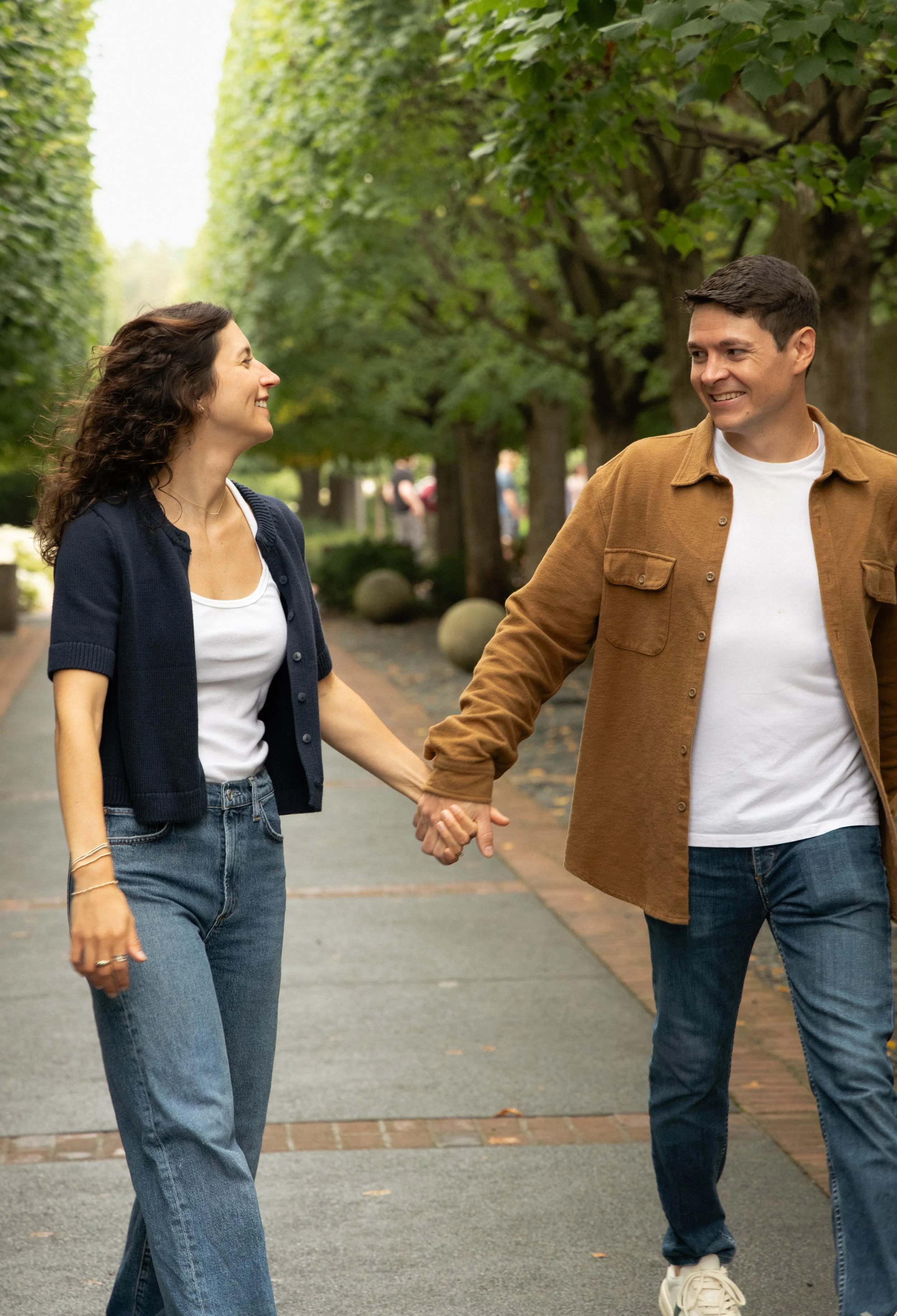 A couple walking in a park, holding hands and smiling at each other, with trees lining the pathway.