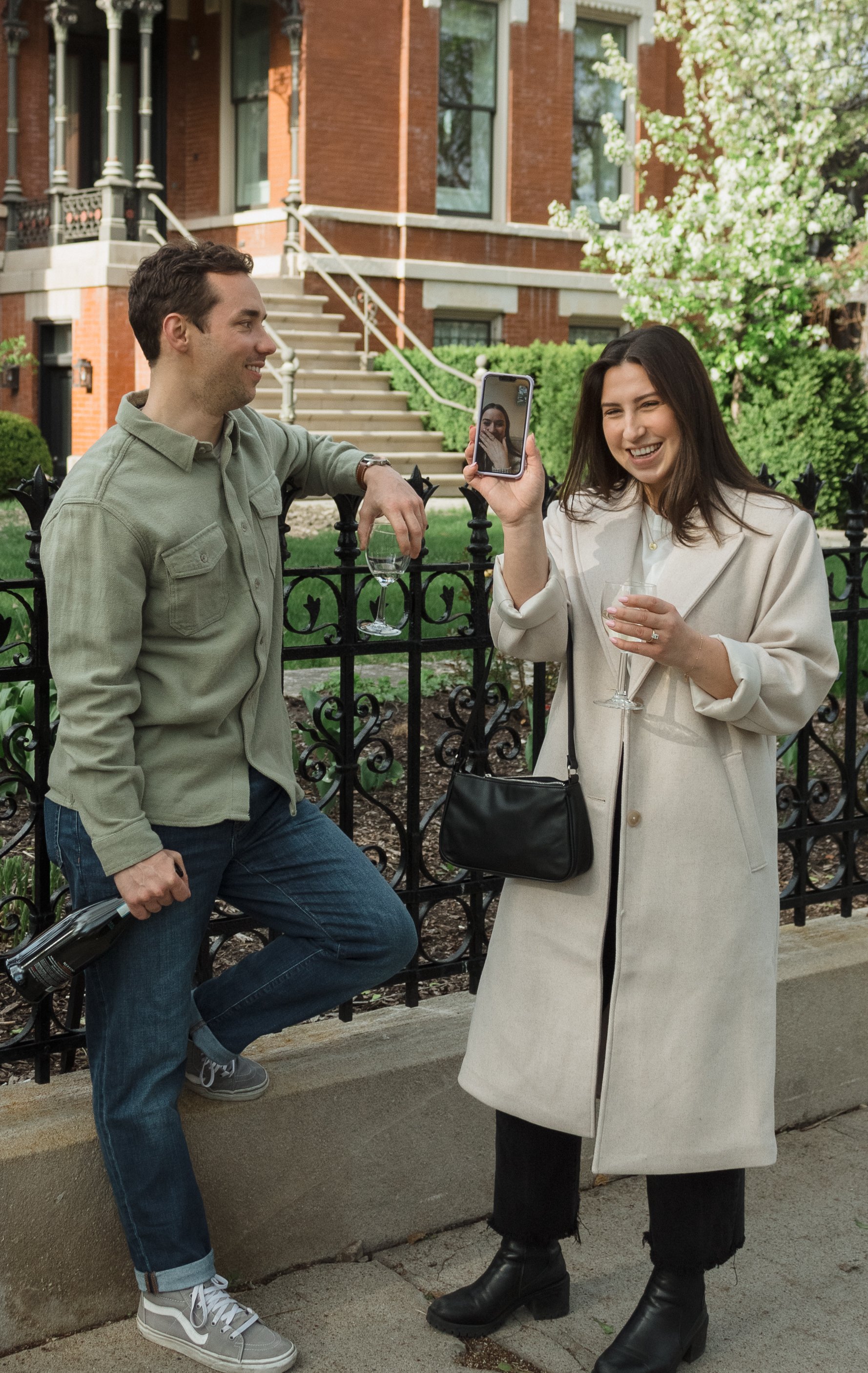 A man and woman are outdoors, with the woman taking a selfie with her phone, both smiling. The man is leaning on a black fence, holding a wine glass and a bottle, while the woman is also holding a wine glass and standing beside him.