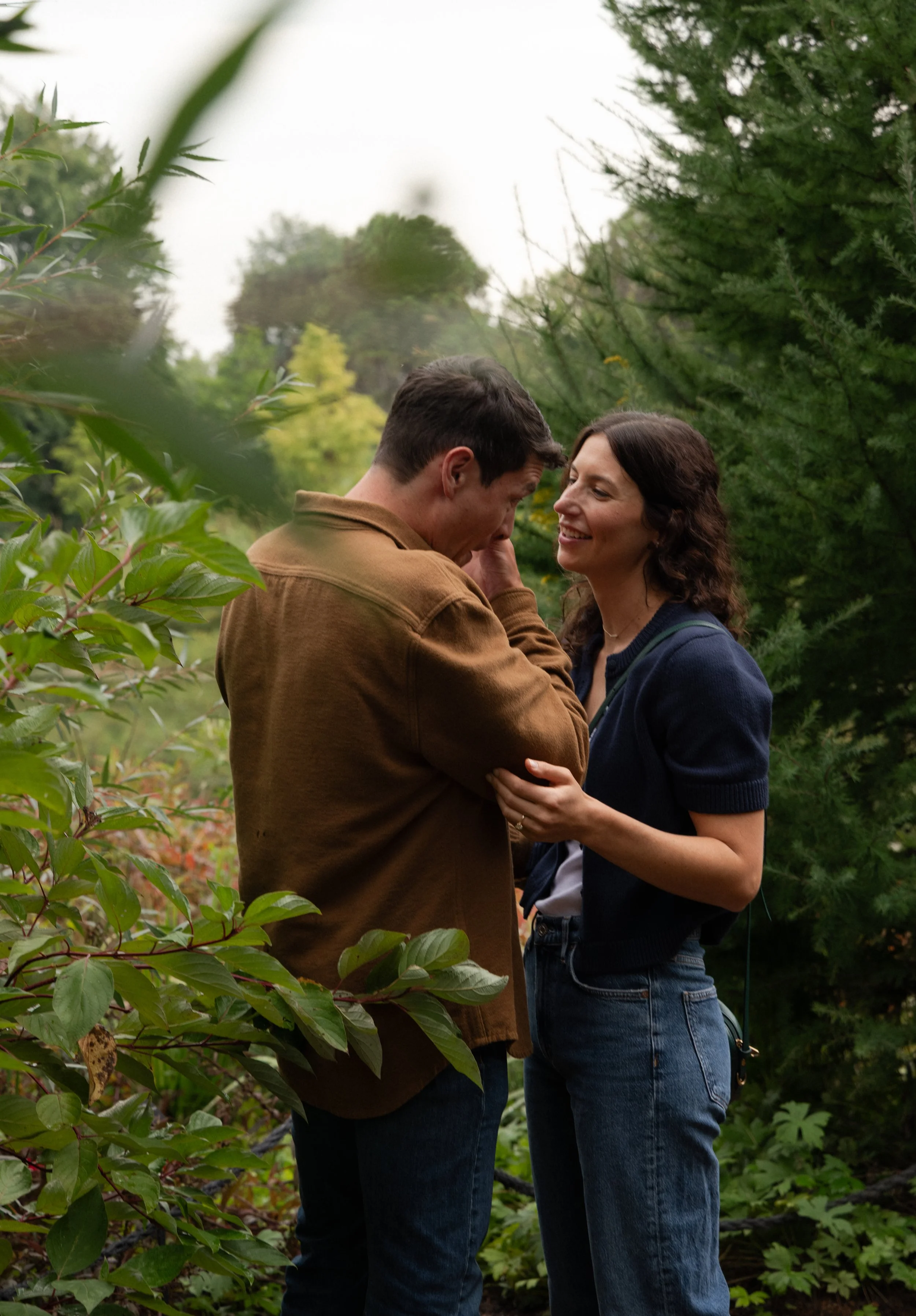 A man and woman stand close together outdoors among trees and bushes. The man is smiling, wearing a brown jacket. The woman is smiling, wearing a dark blue cardigan and jeans. They are holding hands and looking at each other affectionately.