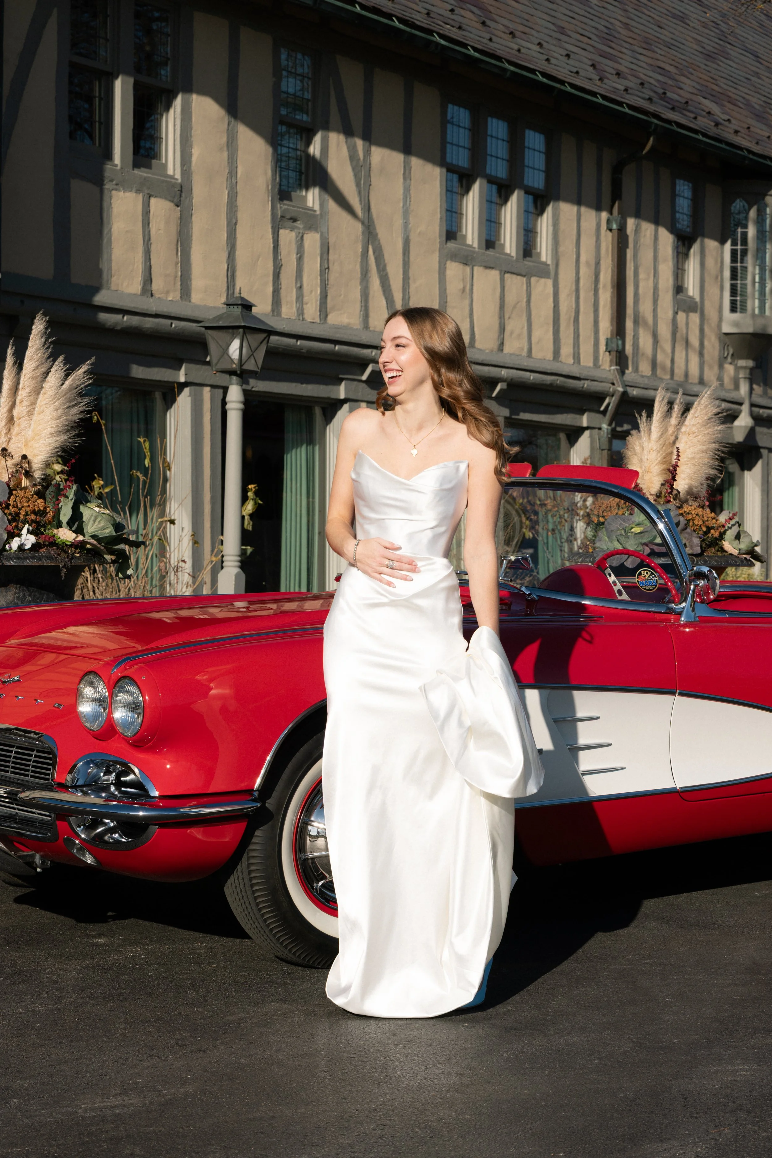 A woman in a white wedding dress standing next to a red vintage convertible car, smiling outdoors in front of a Tudor-style building with floral arrangements.