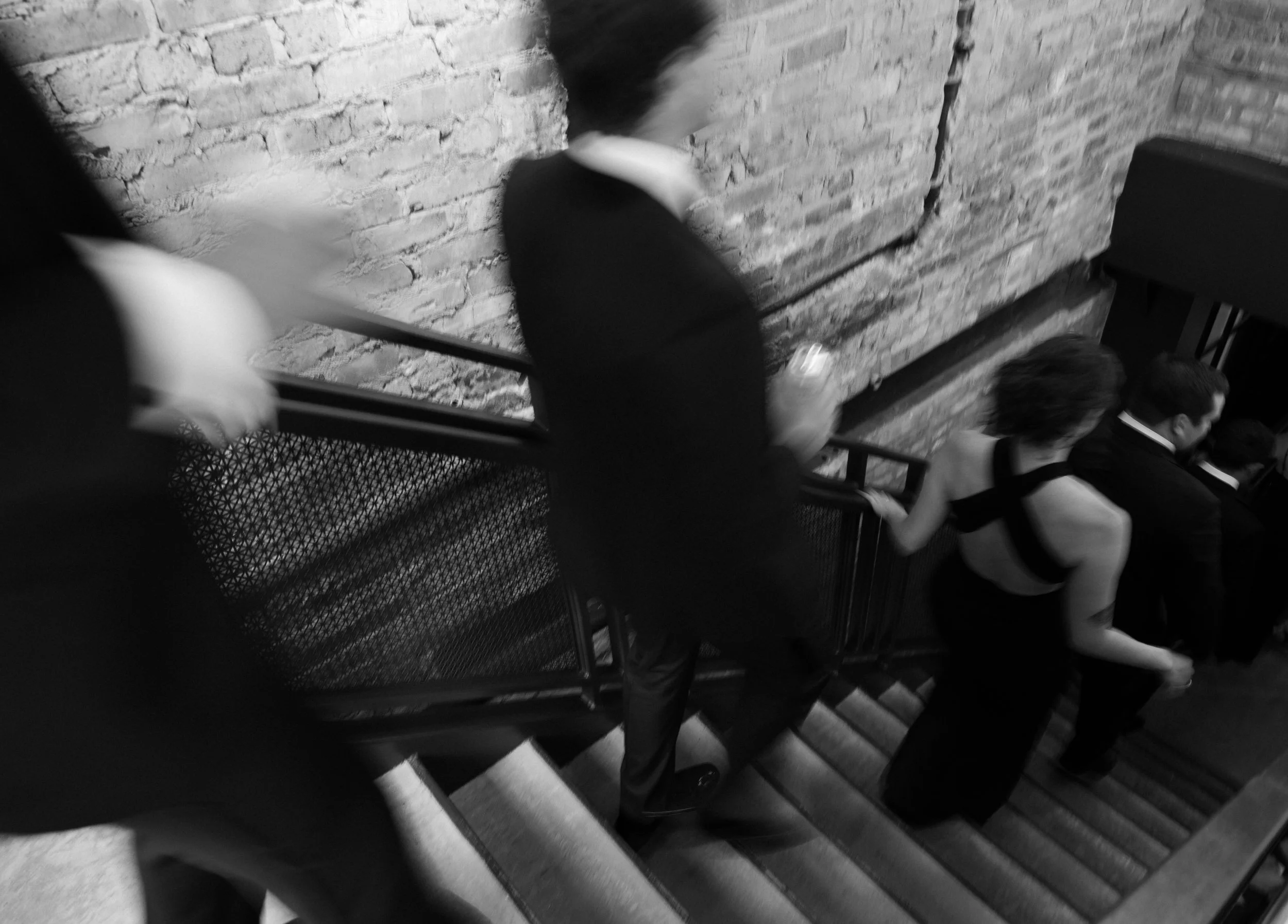 Black-and-white photo of people climbing a staircase. Some individuals are dressed in formal attire, while others are wearing more elegant outfits, suggesting a social event or gathering.