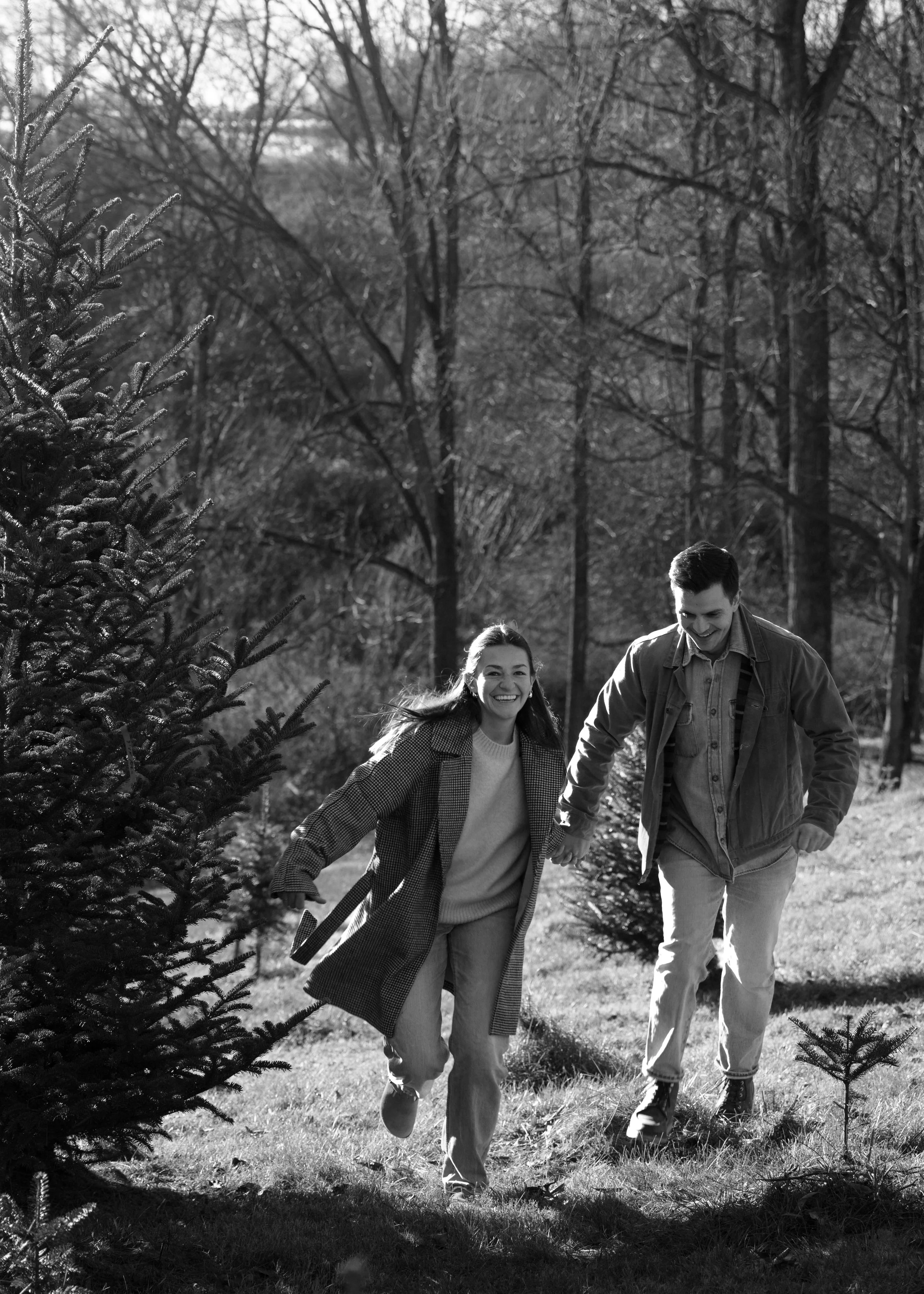 A smiling woman and man walking hand-in-hand through a park with small trees, surrounded by leafless trees in the background, in black and white.