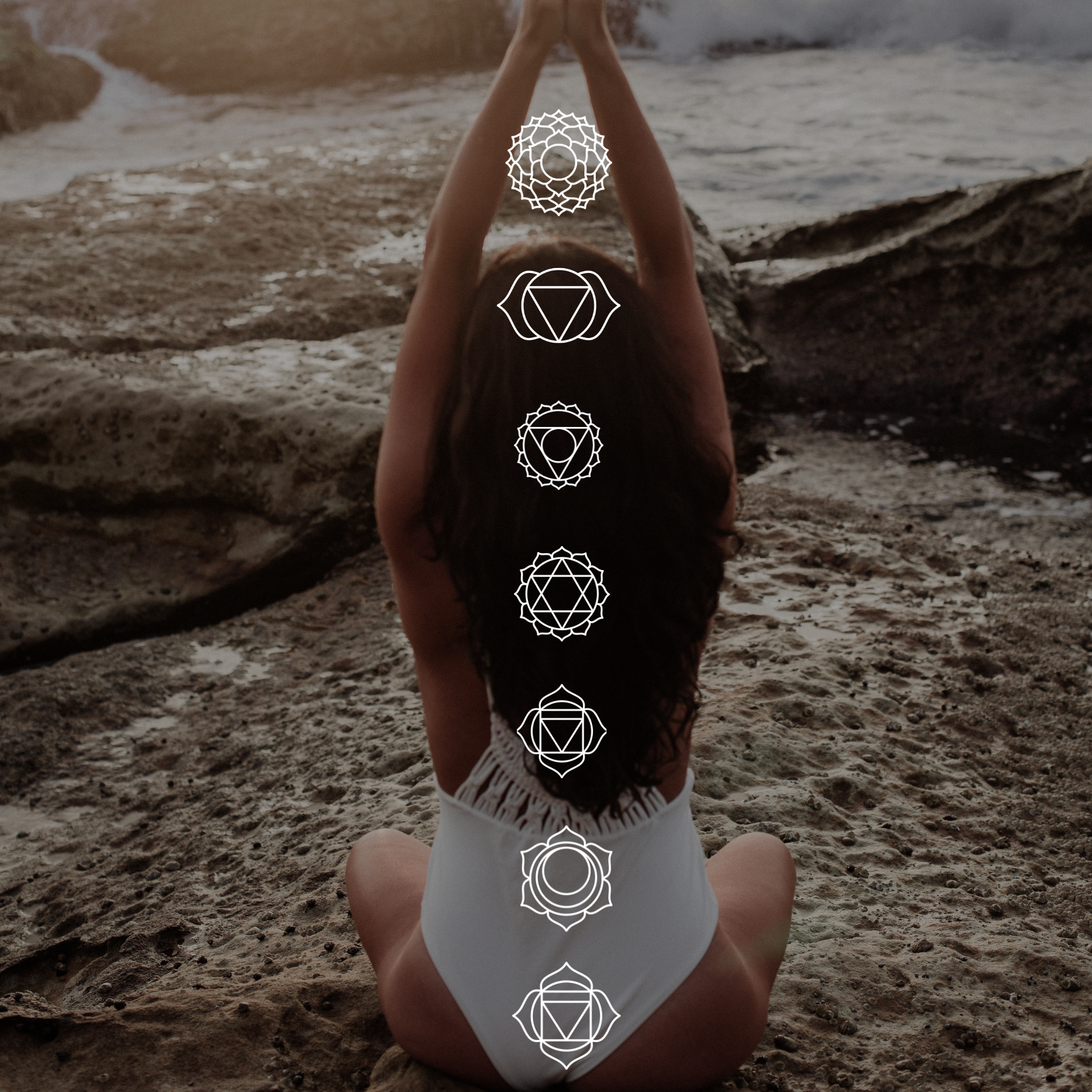 Woman seated on a rocky beach in a white swimsuit, practicing yoga with hands raised above her head. Overlay of chakra symbols aligned along her spine, with a distant ocean in the background.