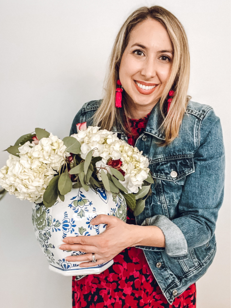 A woman holding a decorative floral vase with white hydrangeas and greenery, posing in front of a plain white wall.