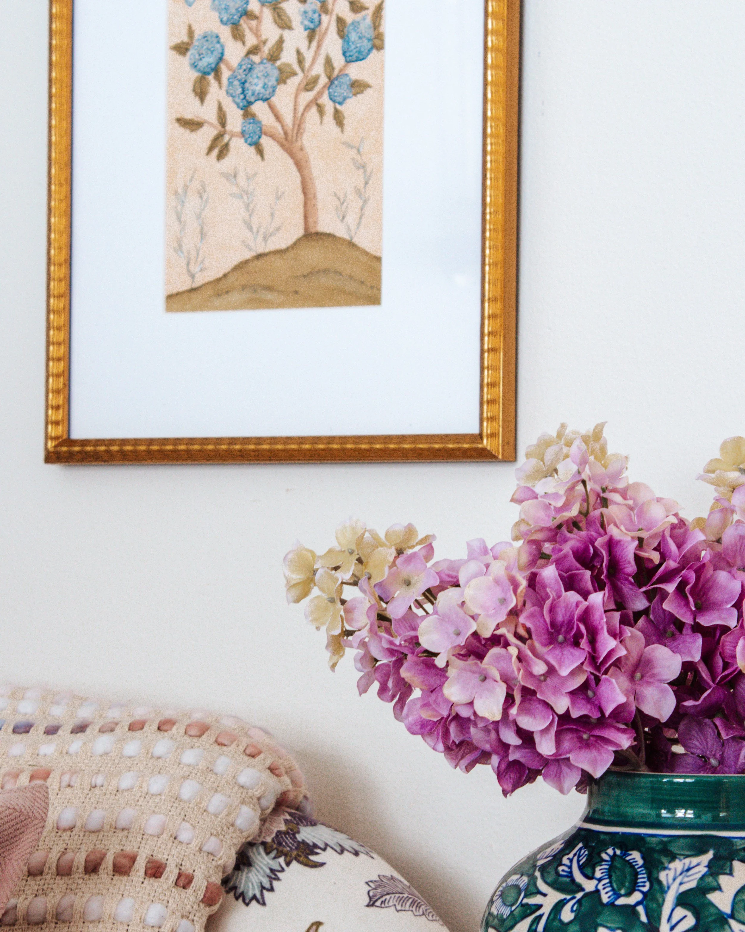 A framed picture of a tree with blue flowers hangs on a white wall above a bouquet of purple and cream hydrangeas in a patterned vase next to a chair with a woven blanket.