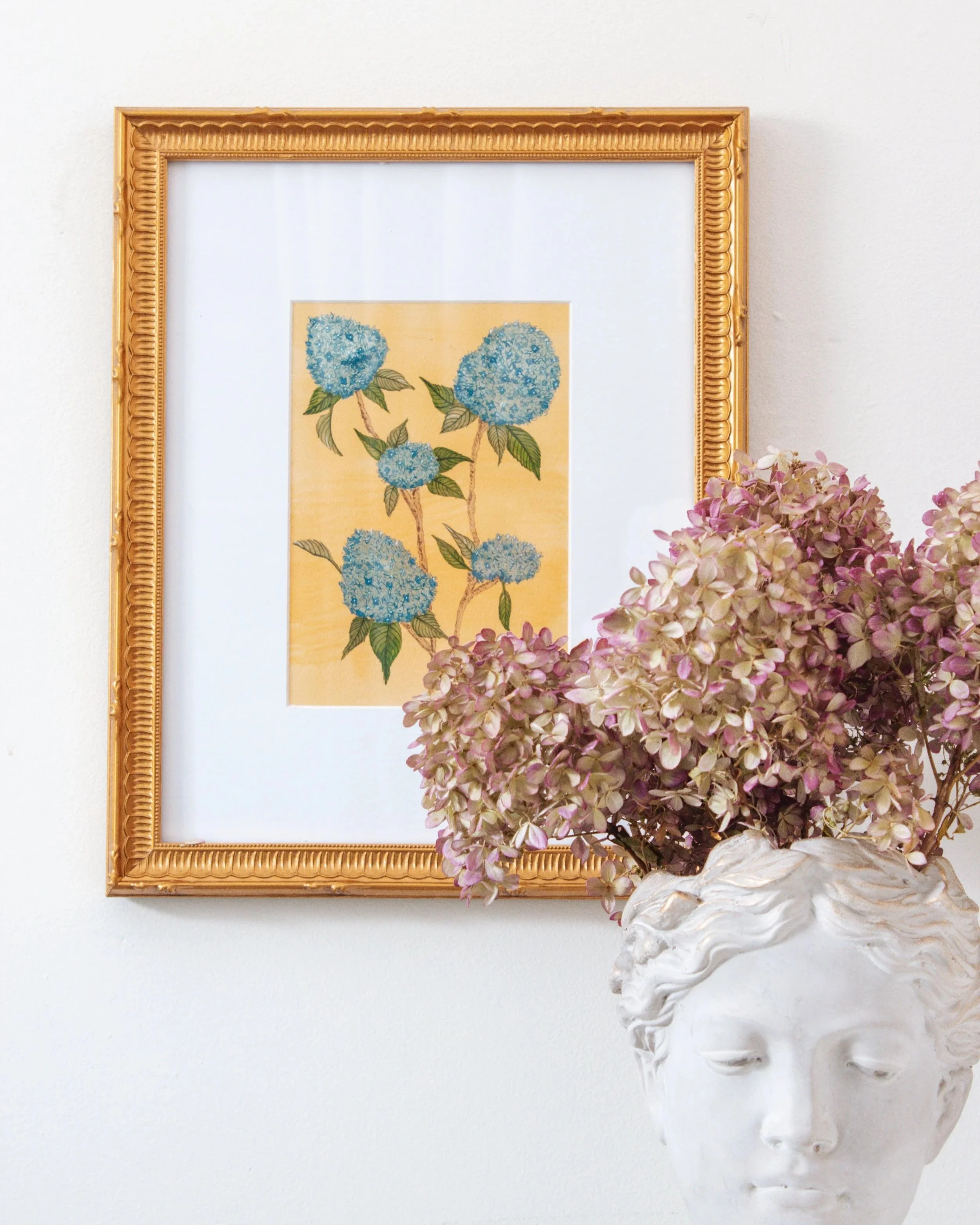 A white wall with a gold framed picture of blue hydrangea flowers hanging next to a sculptural vase with pink hydrangeas.