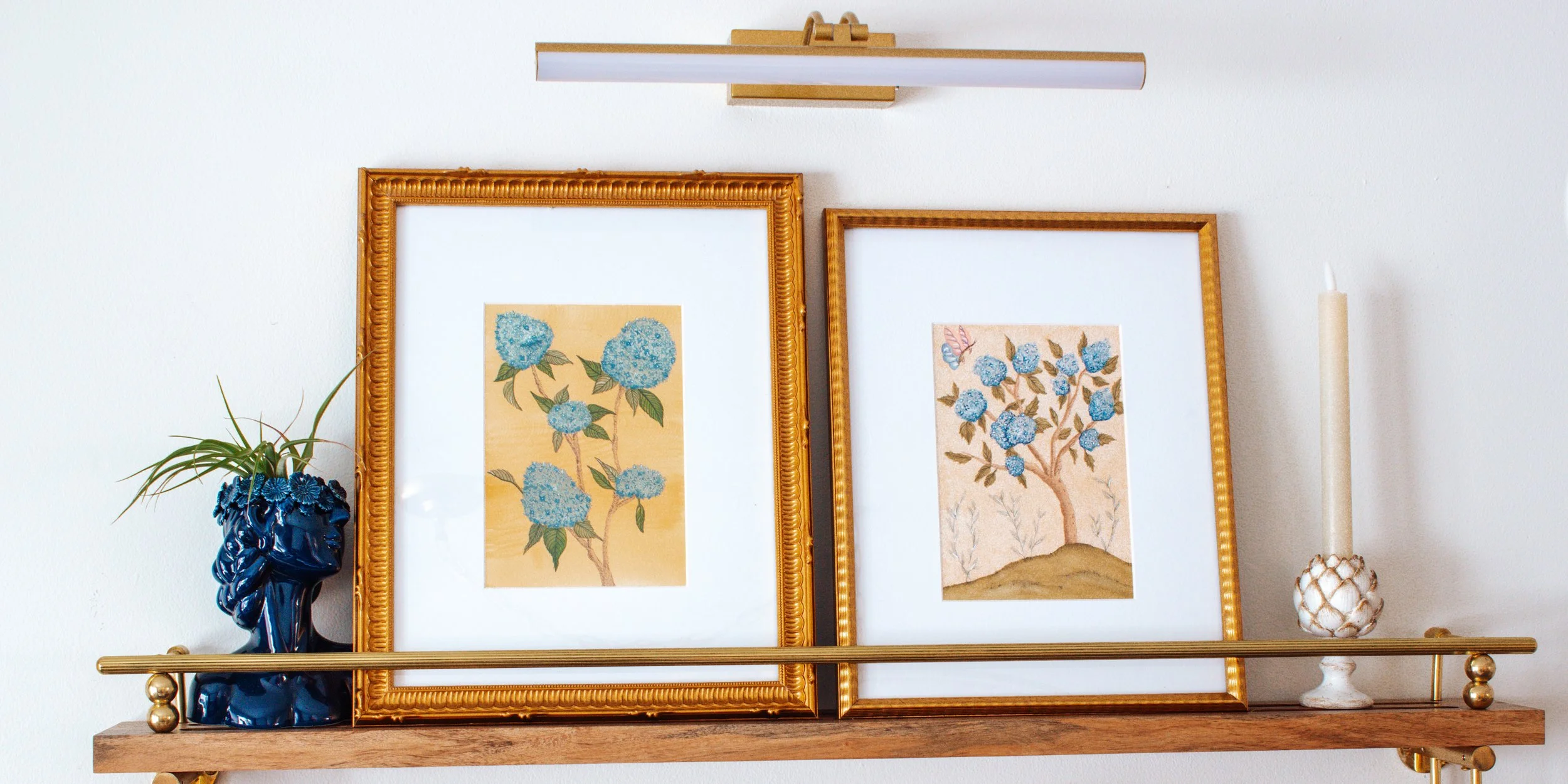 Decorative wall shelf holding two framed botanical prints, a blue ceramic vase with plant, and a taper candle in a white floral holder.