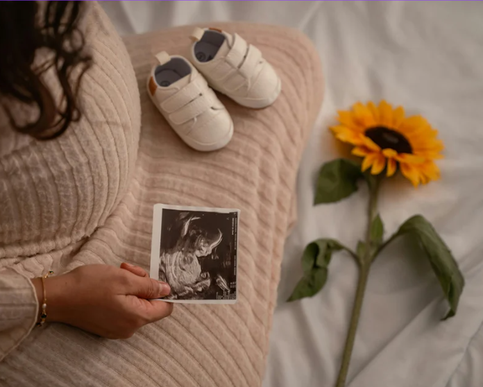 Pregnant woman sitting on bed holding ultrasound photo, with baby shoes nearby and a sunflower on the bed.