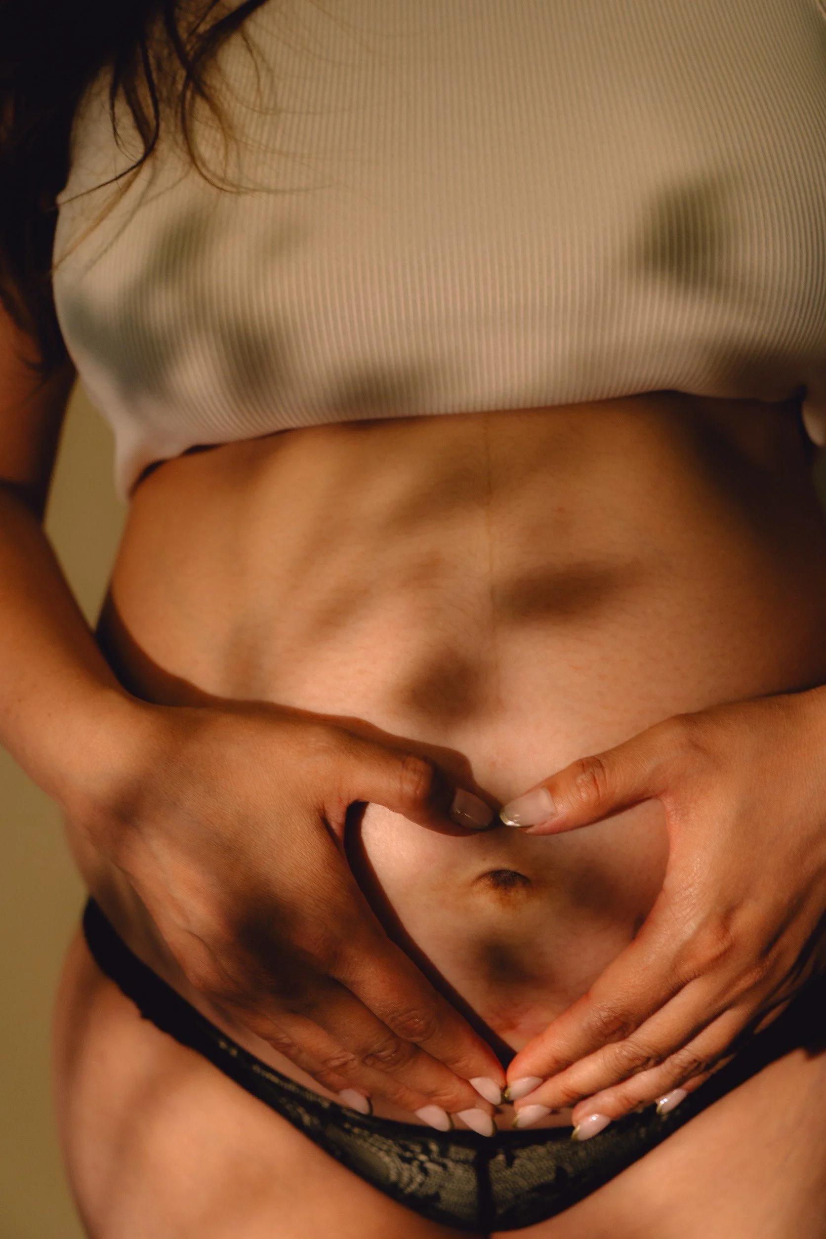 Close-up of a person's torso and hands forming a heart shape around their navel, wearing black lace underwear and a beige top.