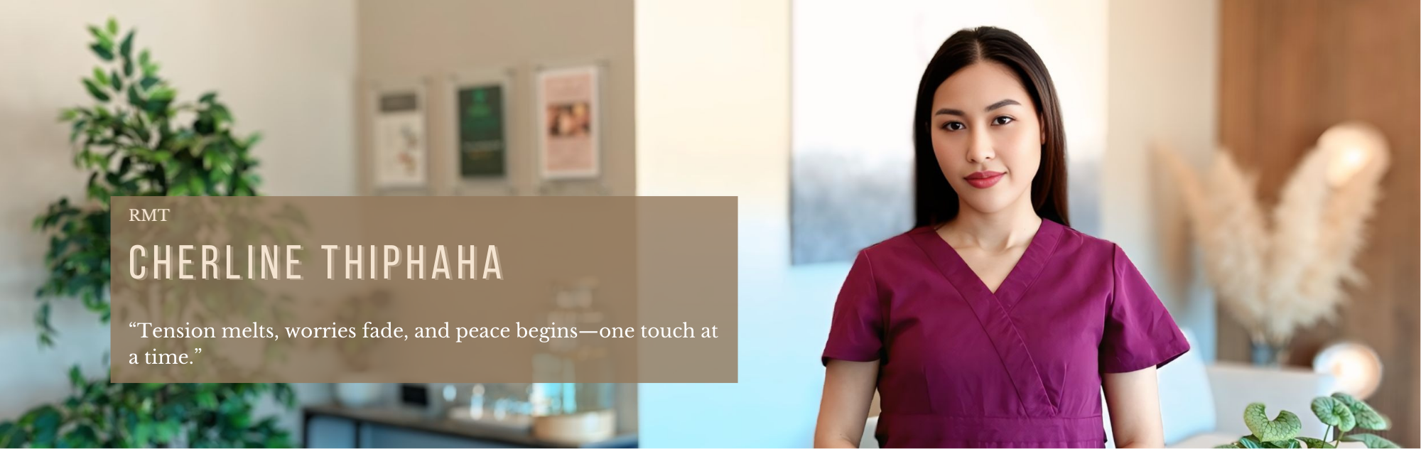 A woman in a purple scrubs standing indoors with a plant and framed pictures in the background.
