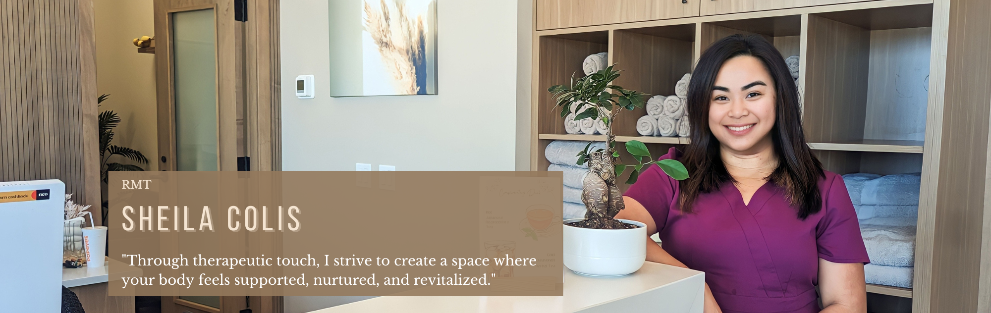 A smiling woman in purple scrubs sitting at a counter with a potted plant, in a cozy, well-decorated room with storing shelves and rolled towels.