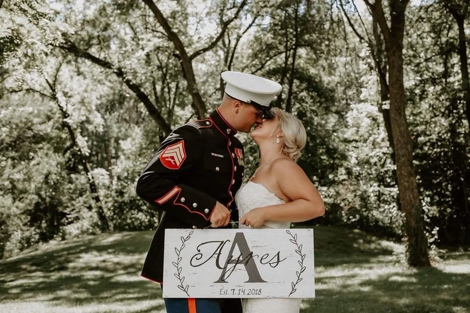 A military couple sharing a kiss outdoors, with a wedding sign in front of them reading 'Alayes' and established date 7.14.2018, surrounded by trees.