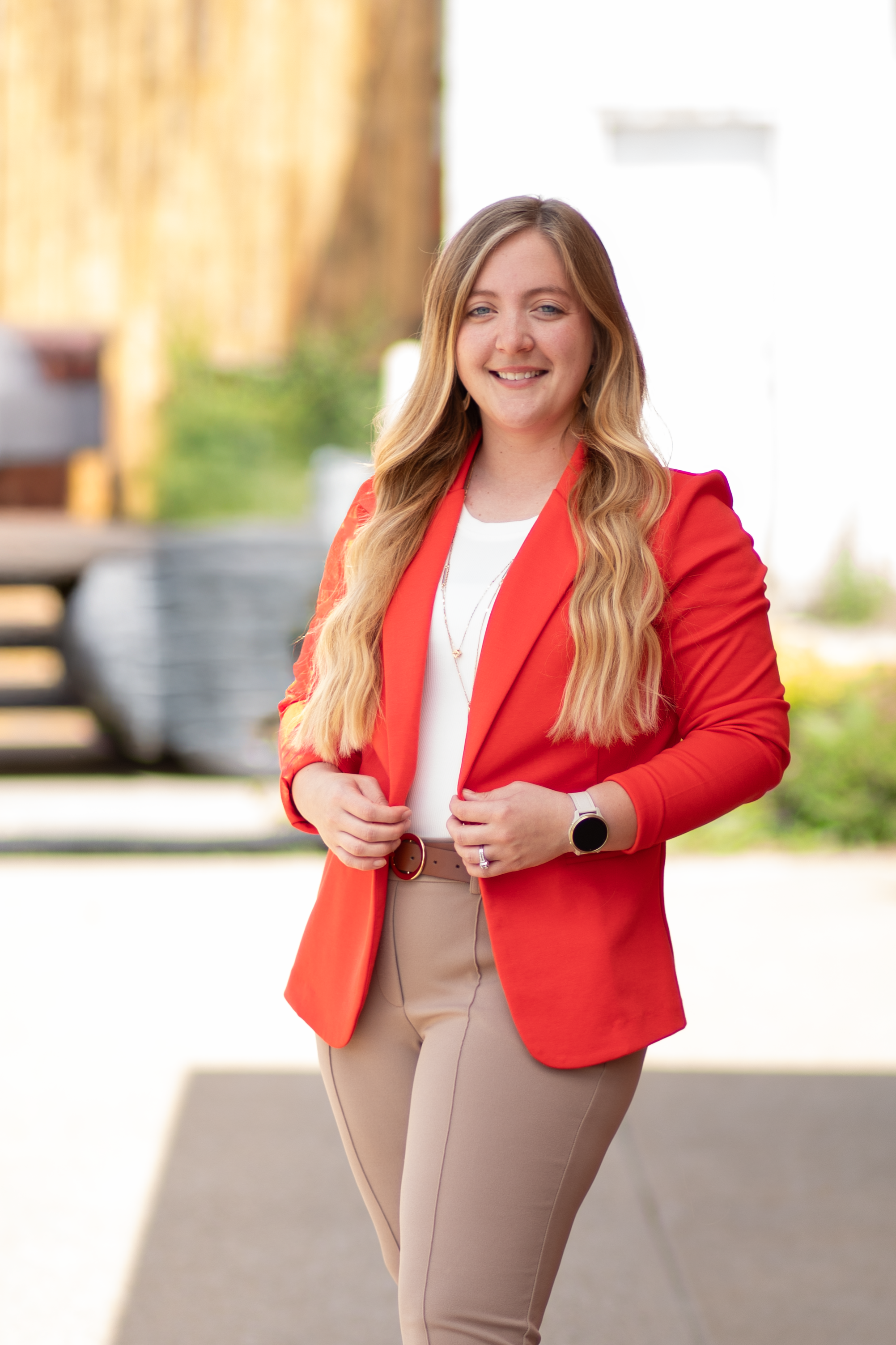 A young woman wearing a red blazer and beige pants standing outdoors with a blurred background.