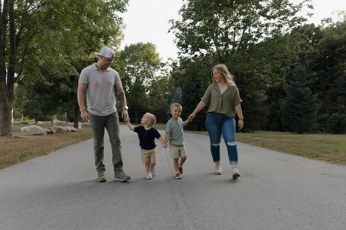 A family of four walking on a park path, holding hands, surrounded by trees.