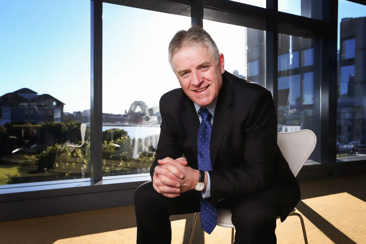 Bob is sitting with a view of the Sydney Harbour behind him, sitting and leaning forward with his elbows on his knees.