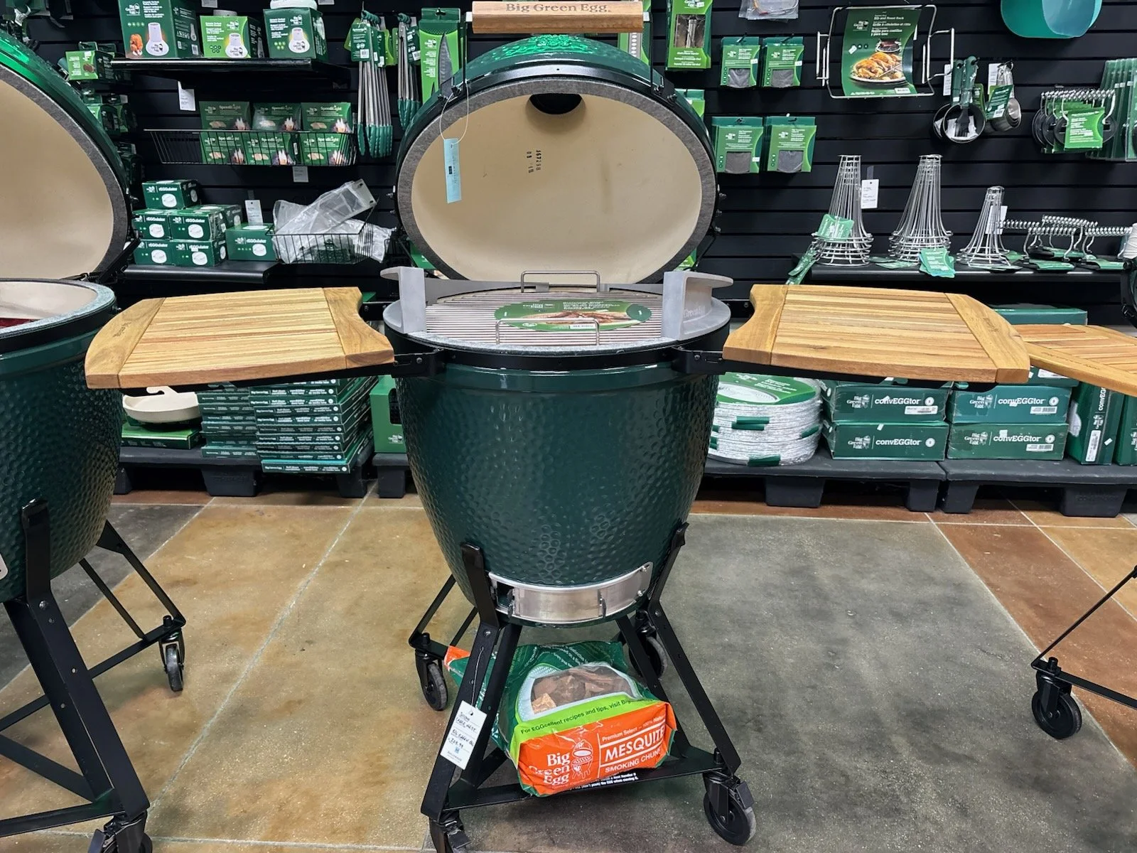A large green outdoor barbecue smoker grill with a wooden side table, in a store aisle with other grilling accessories and accessories on shelves behind it.