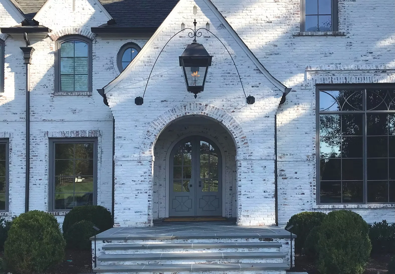 Front view of a white brick house with an arched entryway and steps, large windows, shrubbery in front, and a hanging lantern.