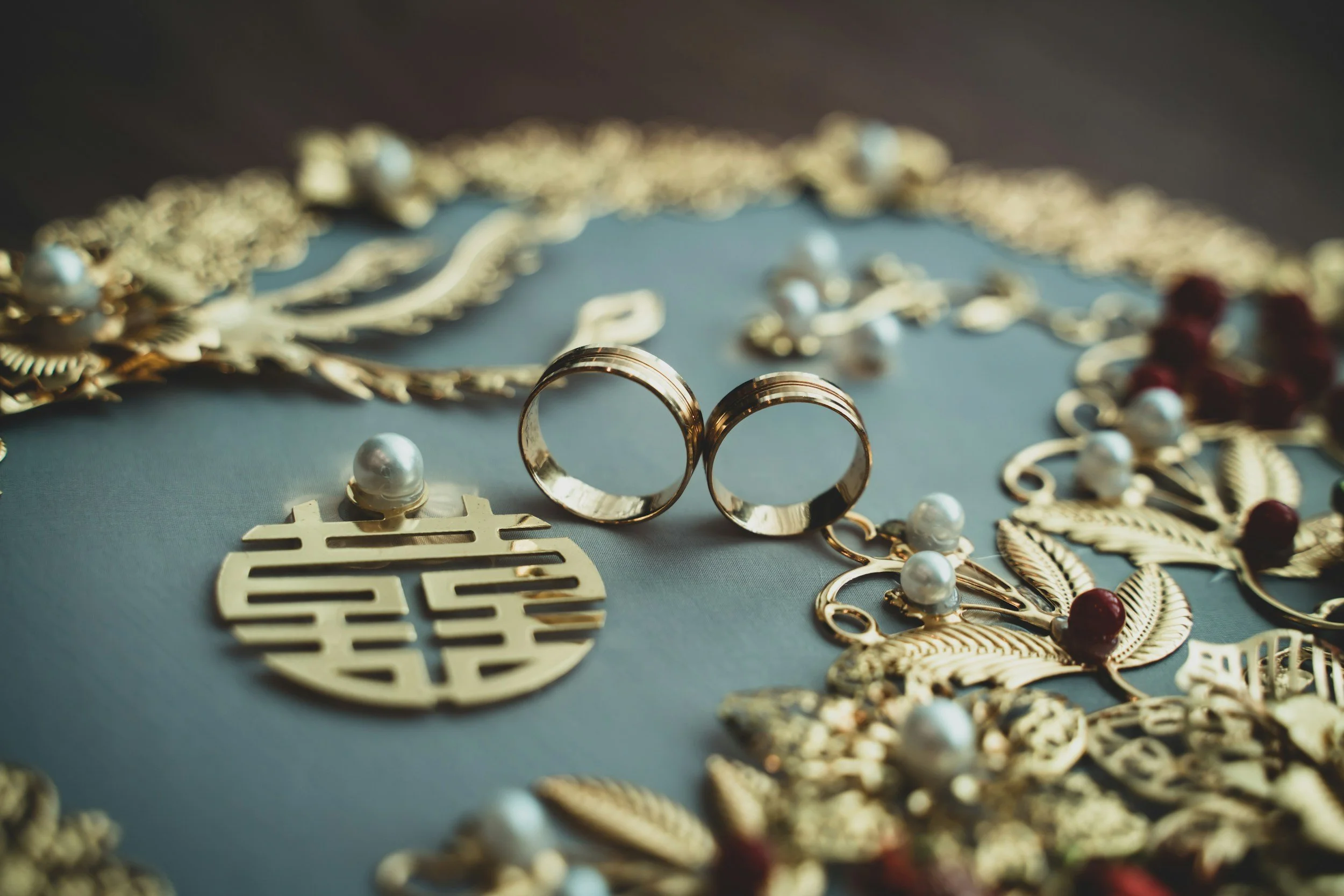 Close-up of two gold wedding bands surrounded by jewelry, pearl necklaces, and a decorative gold pendant with a pearl on top. The jewelry is arranged on a dark surface.