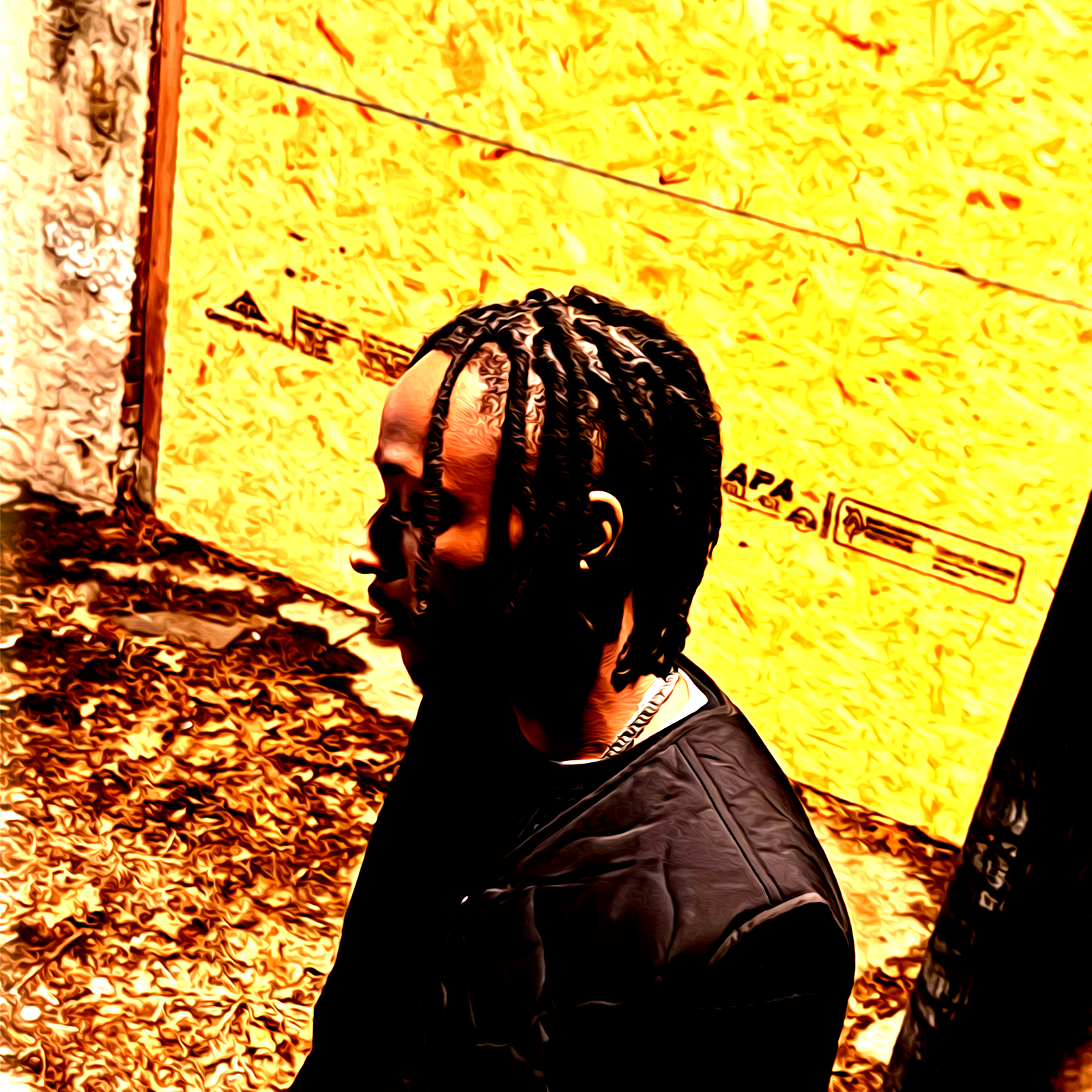 A man with braids and glasses is standing outdoors against a yellow wall with some text, wearing a black jacket.