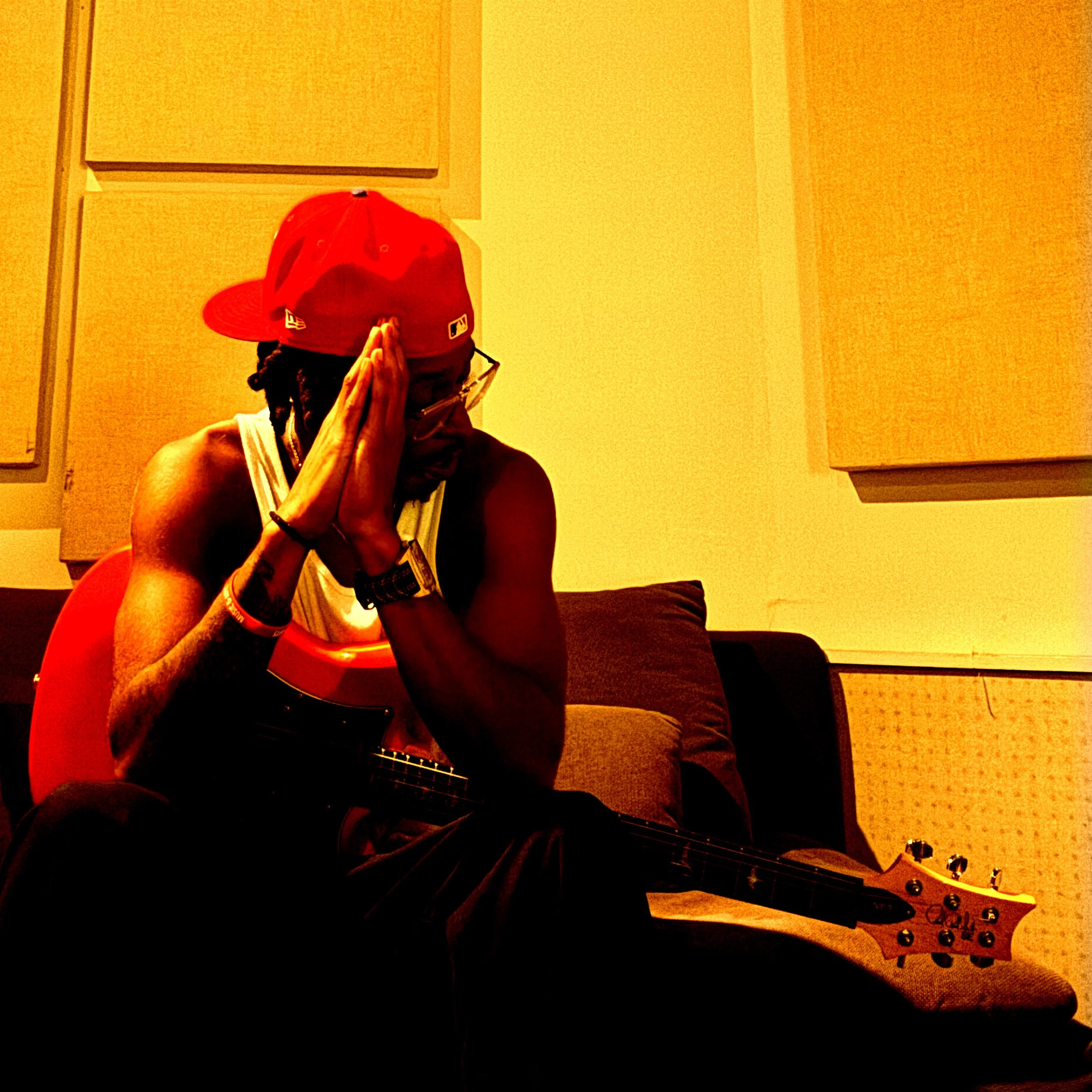 A man sitting on a couch with closed eyes and hands pressed together near his face, holding a guitar on his lap, wearing a red cap, glasses, and a sleeveless shirt, in a room with yellow walls and soundproof panels.