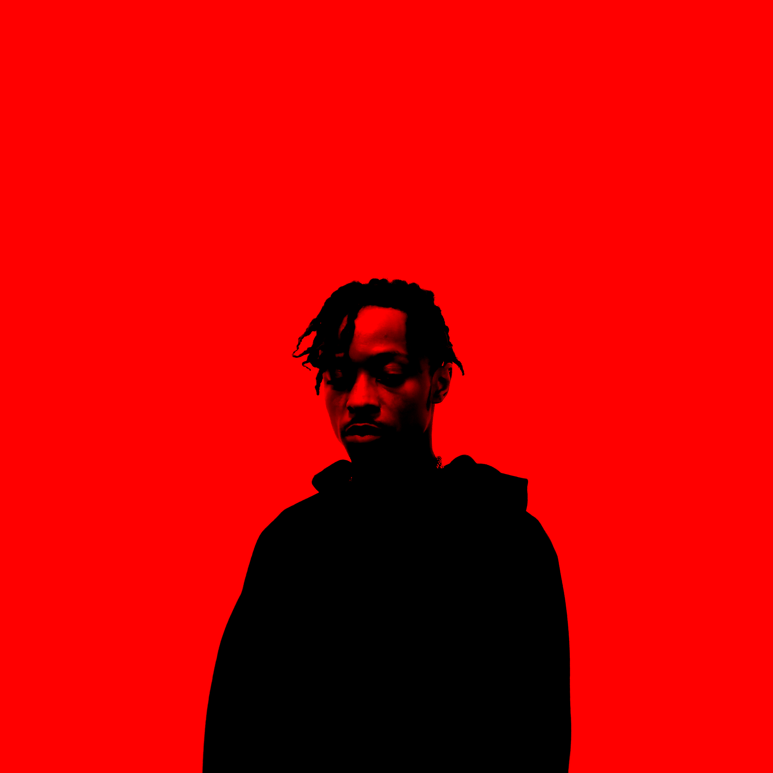 A young man with dreadlocks wearing a black hoodie, looking down, against a bright red background.