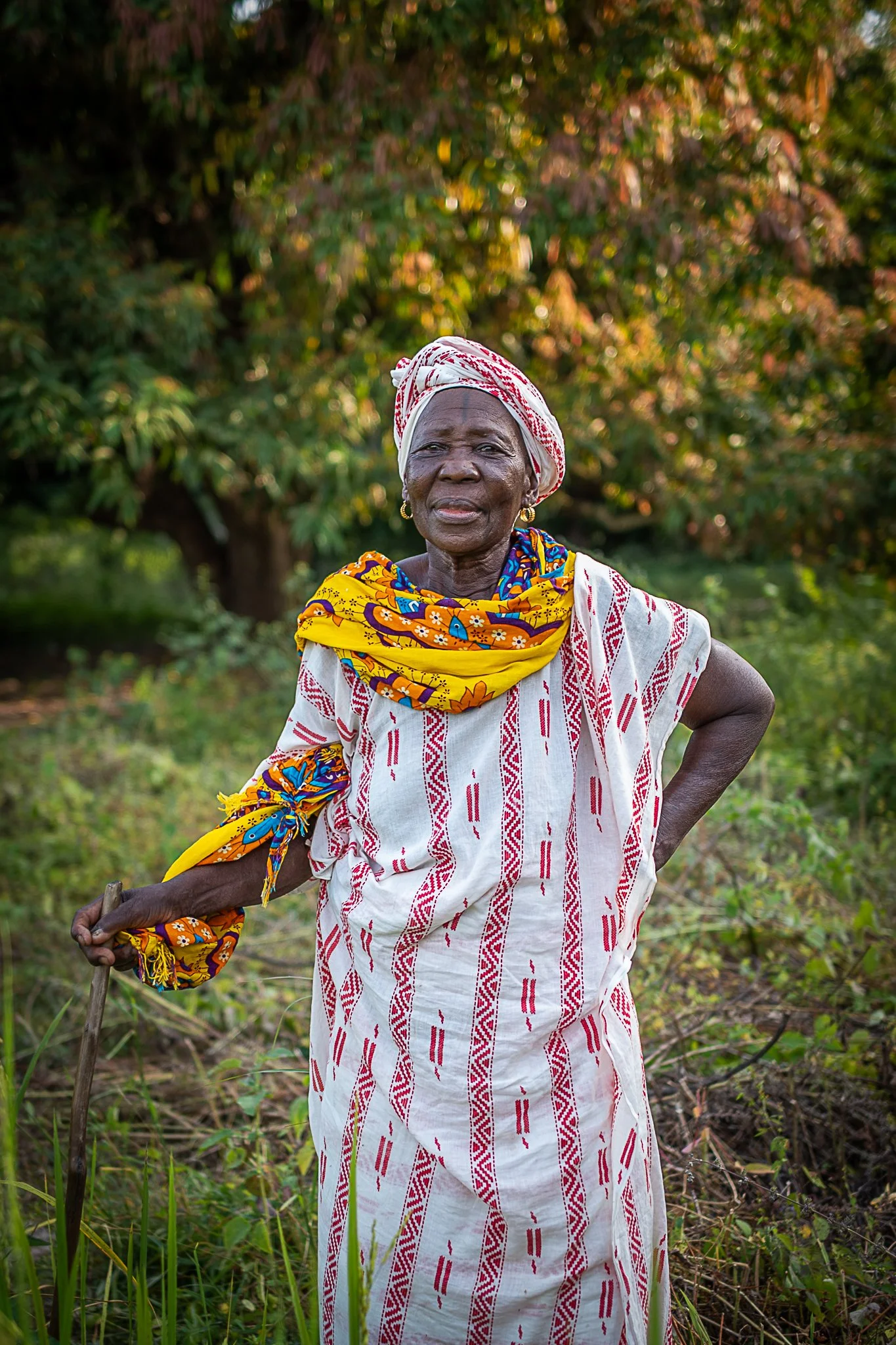An elderly woman wearing traditional African clothing, including a white and red patterned dress, a colorful yellow and blue scarf, and a headwrap, stands outdoors in a grassy area with trees in the background, holding a walking stick and looking at 