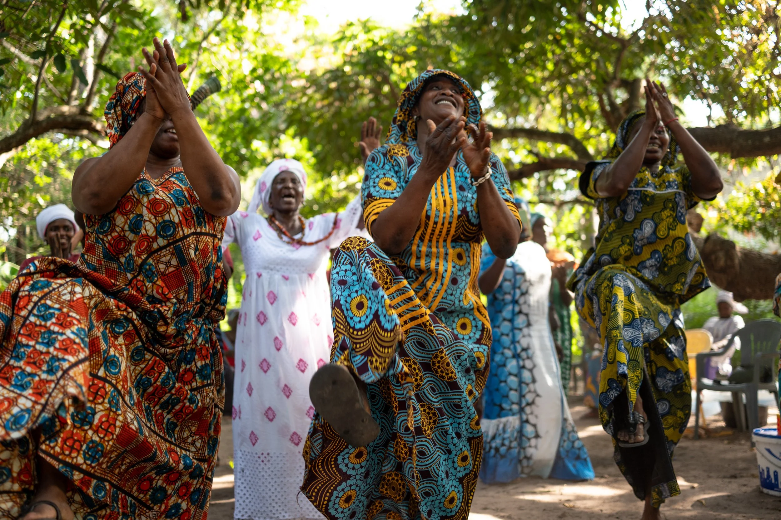 Women dancing and celebrating outdoors under a large tree, wearing colorful traditional dresses.