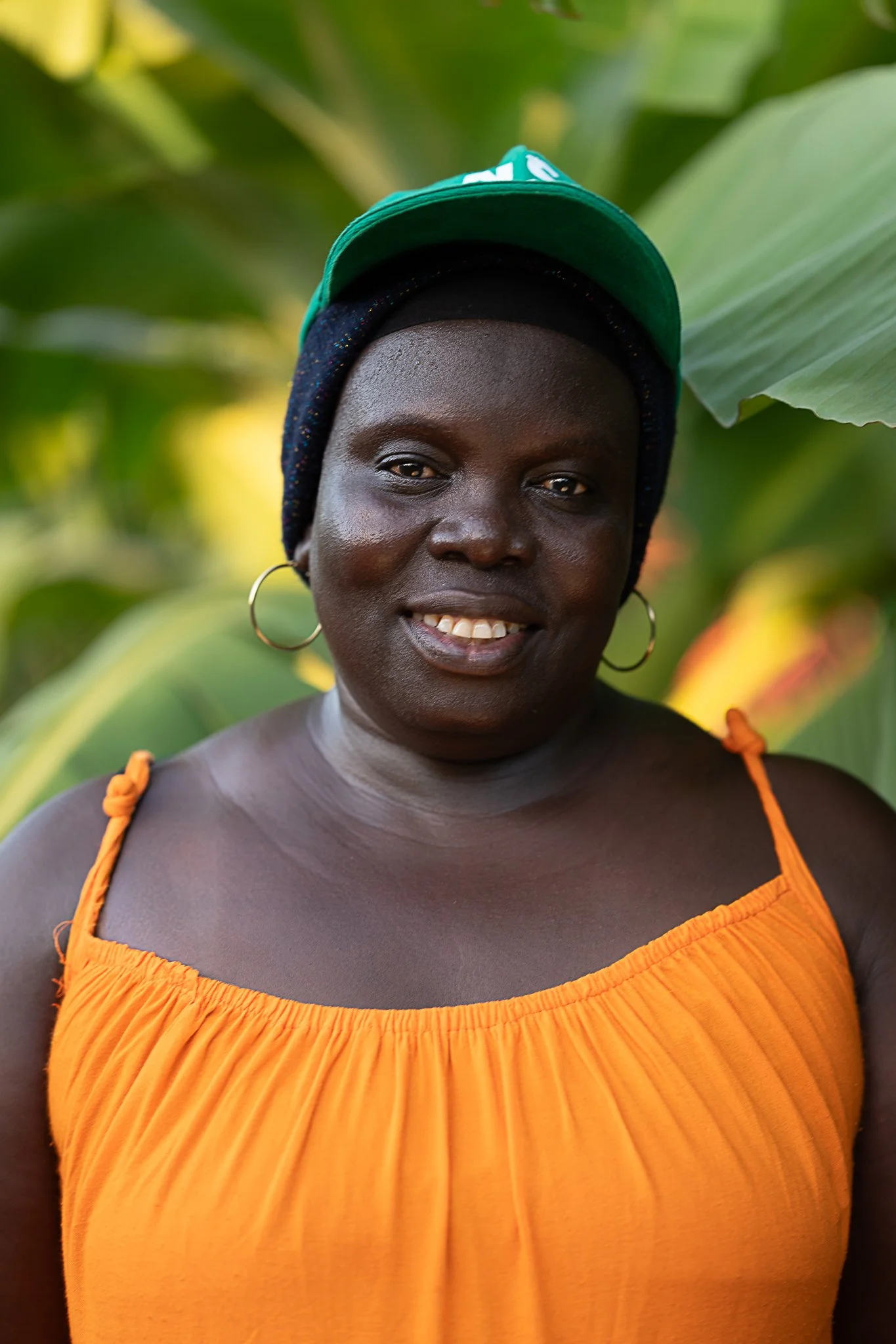 A smiling woman with dark skin, wearing a bright orange sleeveless top, large hoop earrings, a dark beanie with a teal cap on top, and standing outdoors among green leaves.
