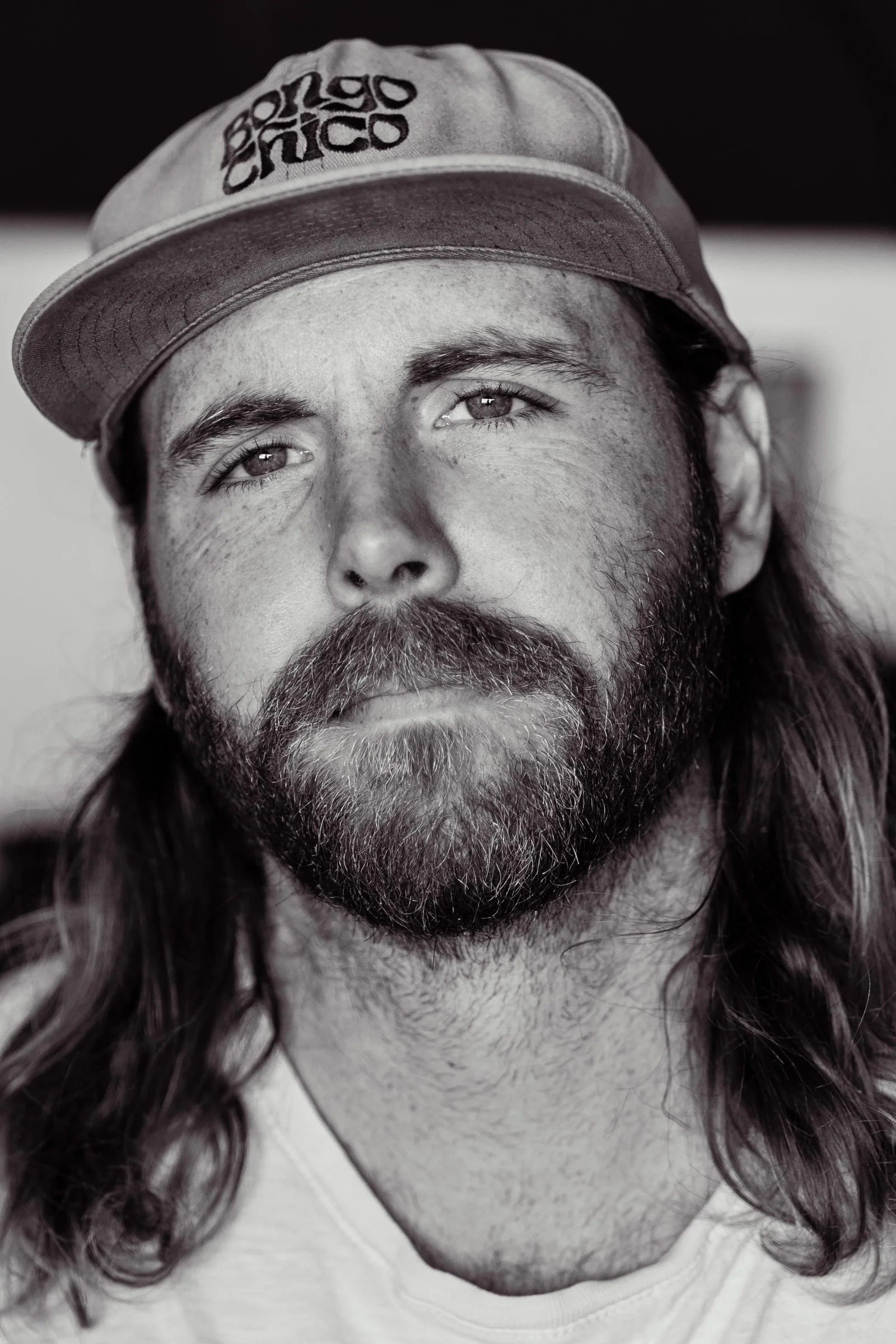 Black and white close-up portrait of a man with long hair, a beard, and mustache, wearing a cap that says 'Poco Chico'.