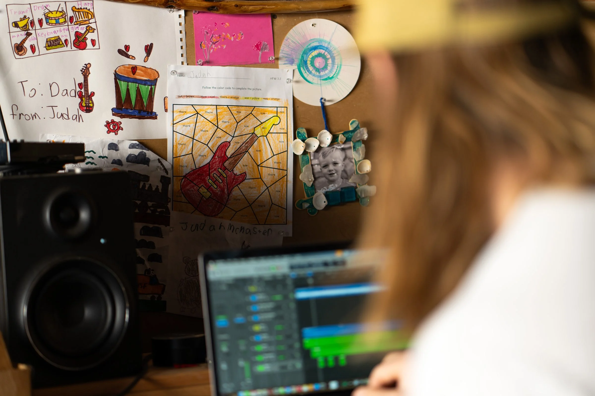 A cluttered desk with children's drawings, a framed photo surrounded by seashells, a computer with audio editing software, and a speaker.