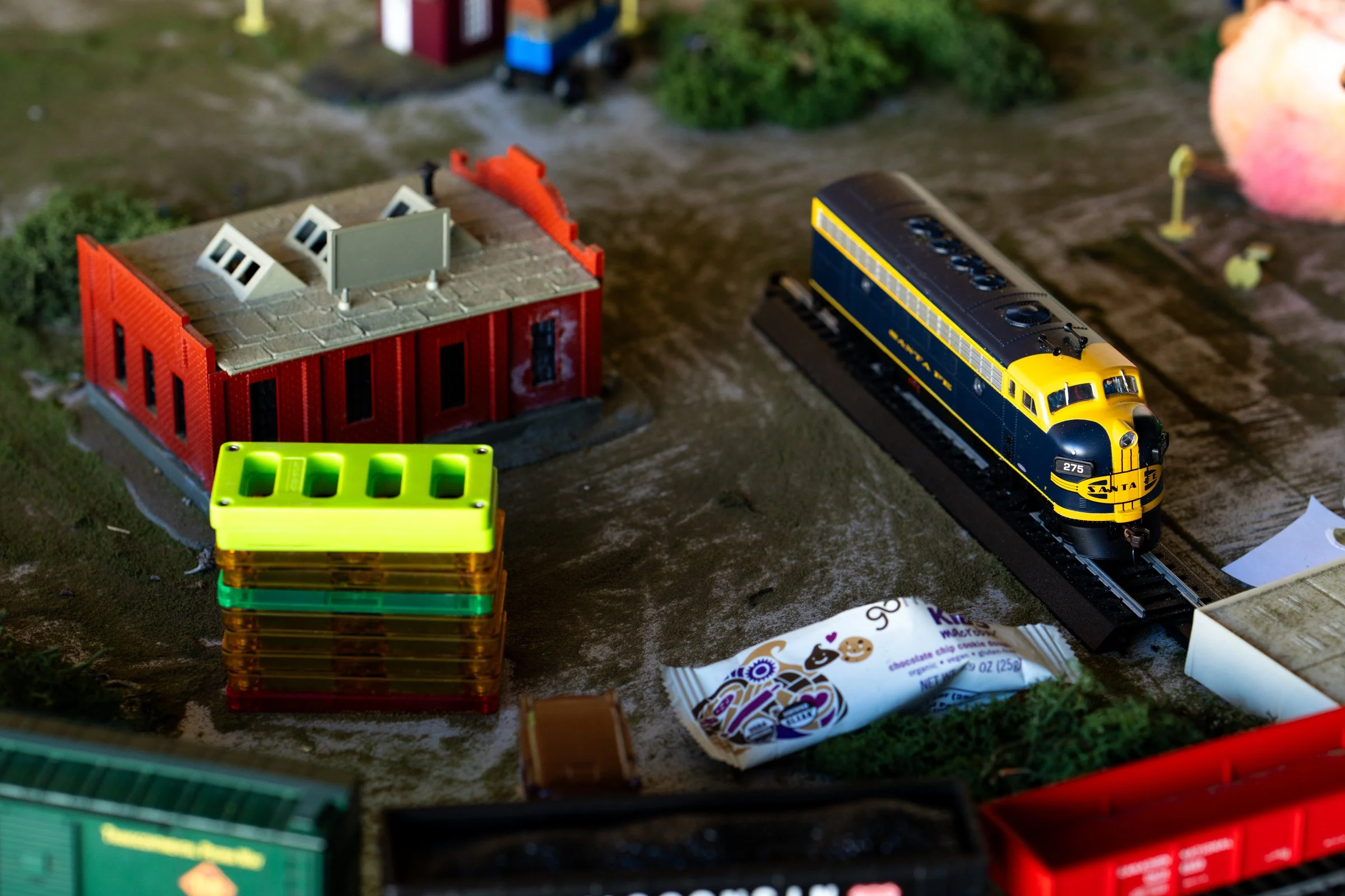 Model train set with a blue and yellow locomotive on tracks, a red building, and various small objects on a miniature landscape.