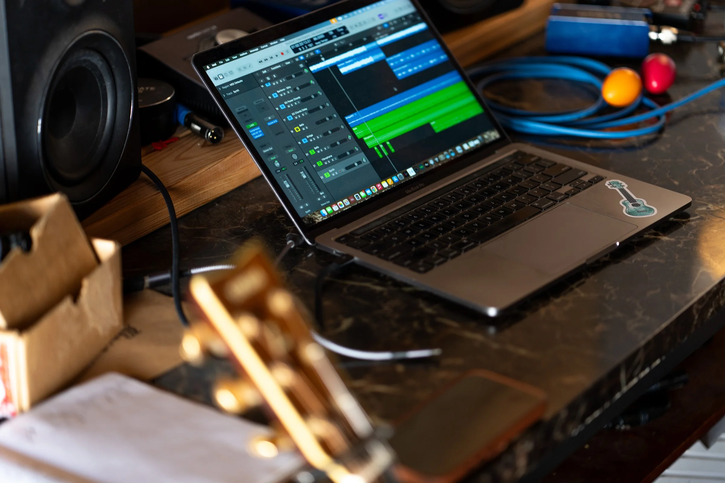 A laptop displaying a music production software on a dark marble table, surrounded by audio equipment, cables, and colorful percussion instruments.