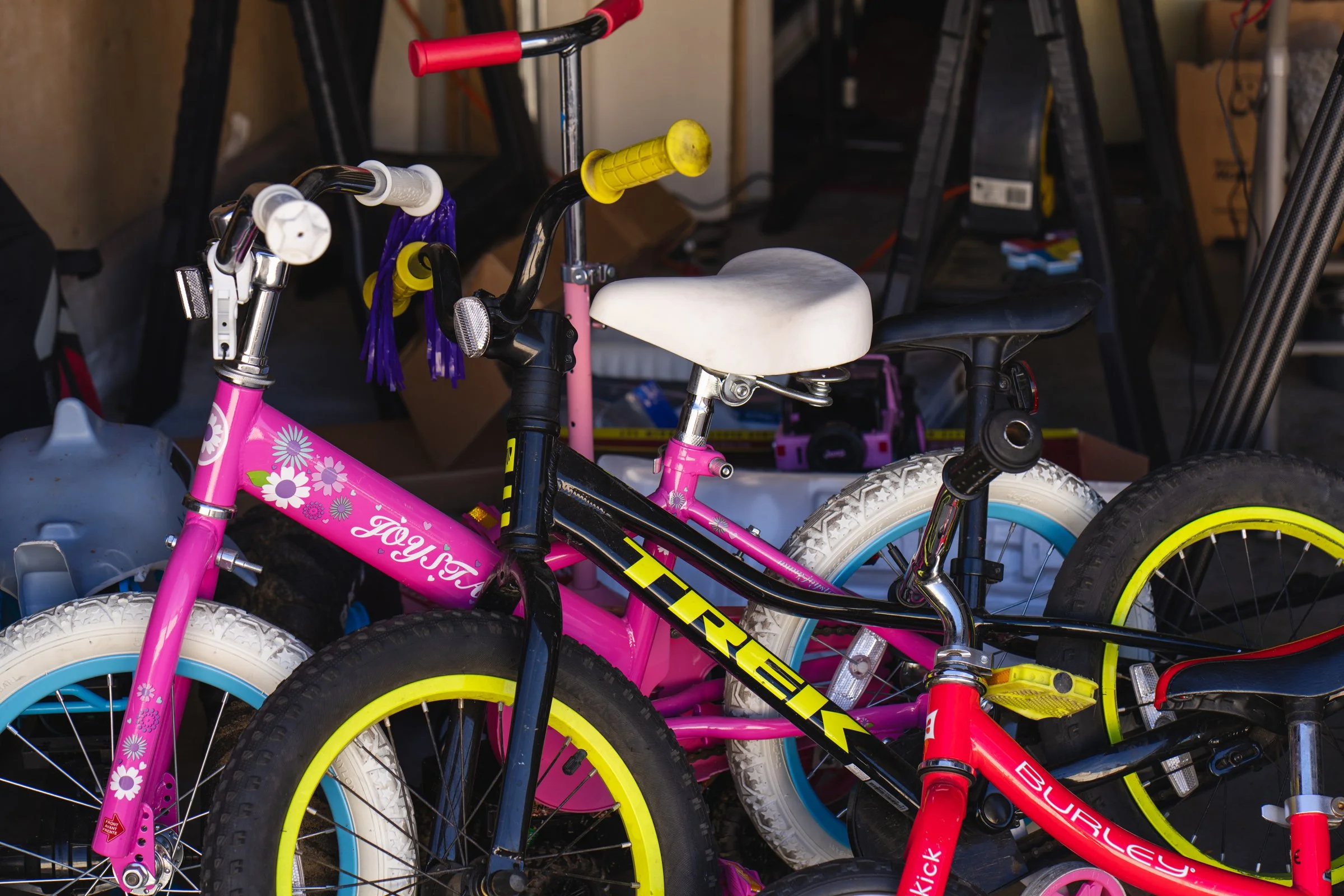 Two children's bicycles, one pink with floral design and the other black with yellow and pink accents, leaning against a cluttered background of a garage or storage space.