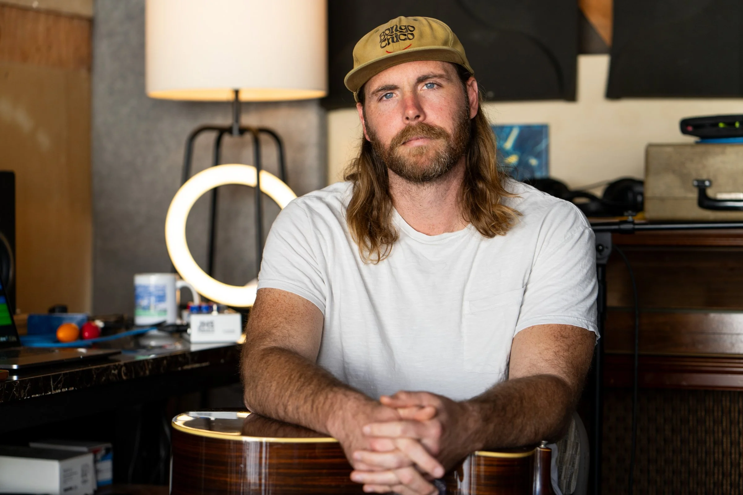 Man with long hair and beard wearing a beige cap and white t-shirt, sitting at a desk with a guitar in front of him, in a room with warm lighting and musical equipment.