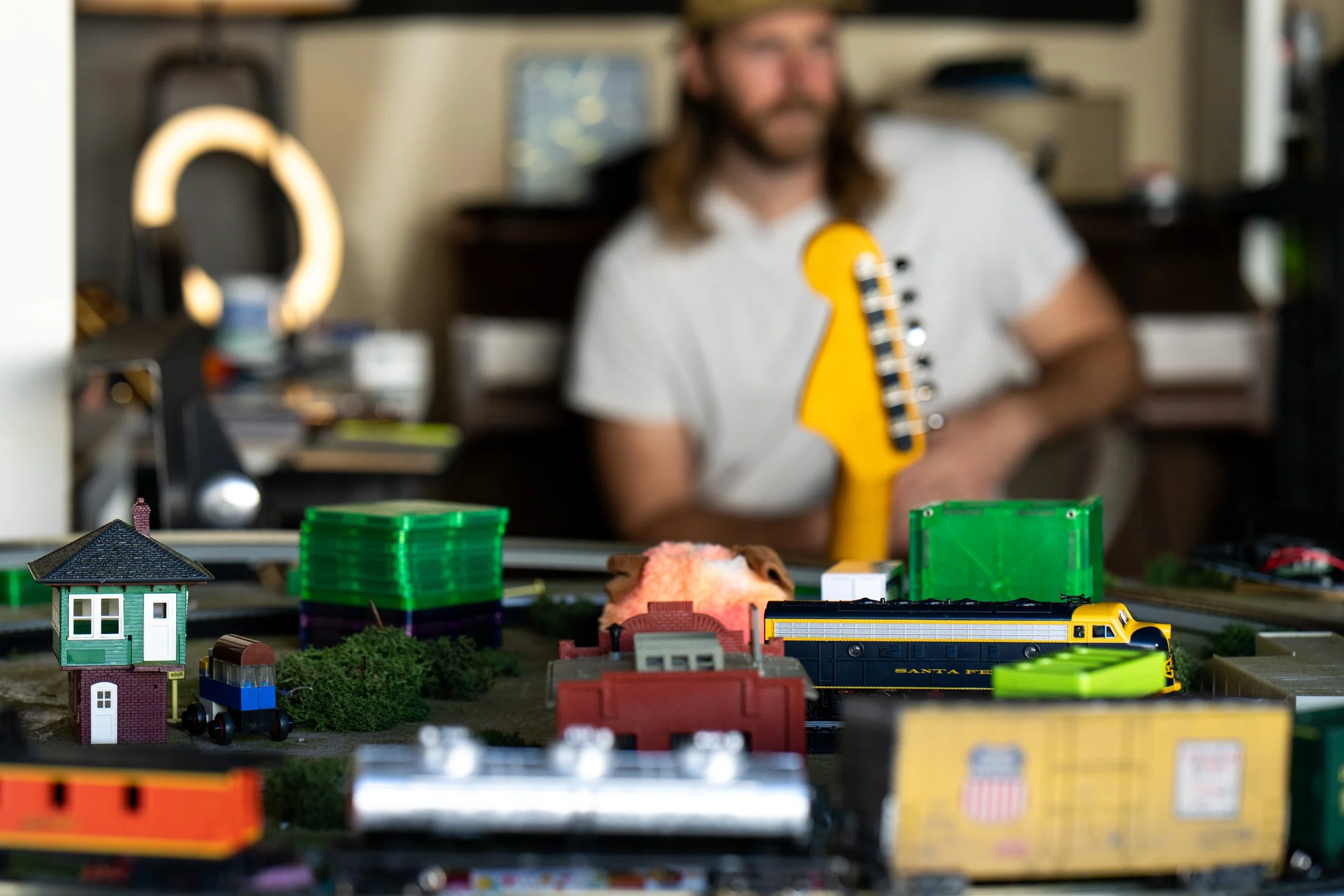 Close-up of a miniature model train set with a yellow and black Santa Fe train, green stackable cargo containers, a small house, and various miniature accessories on a table. A man with long hair and beard, wearing a white shirt, is blurred in the ba