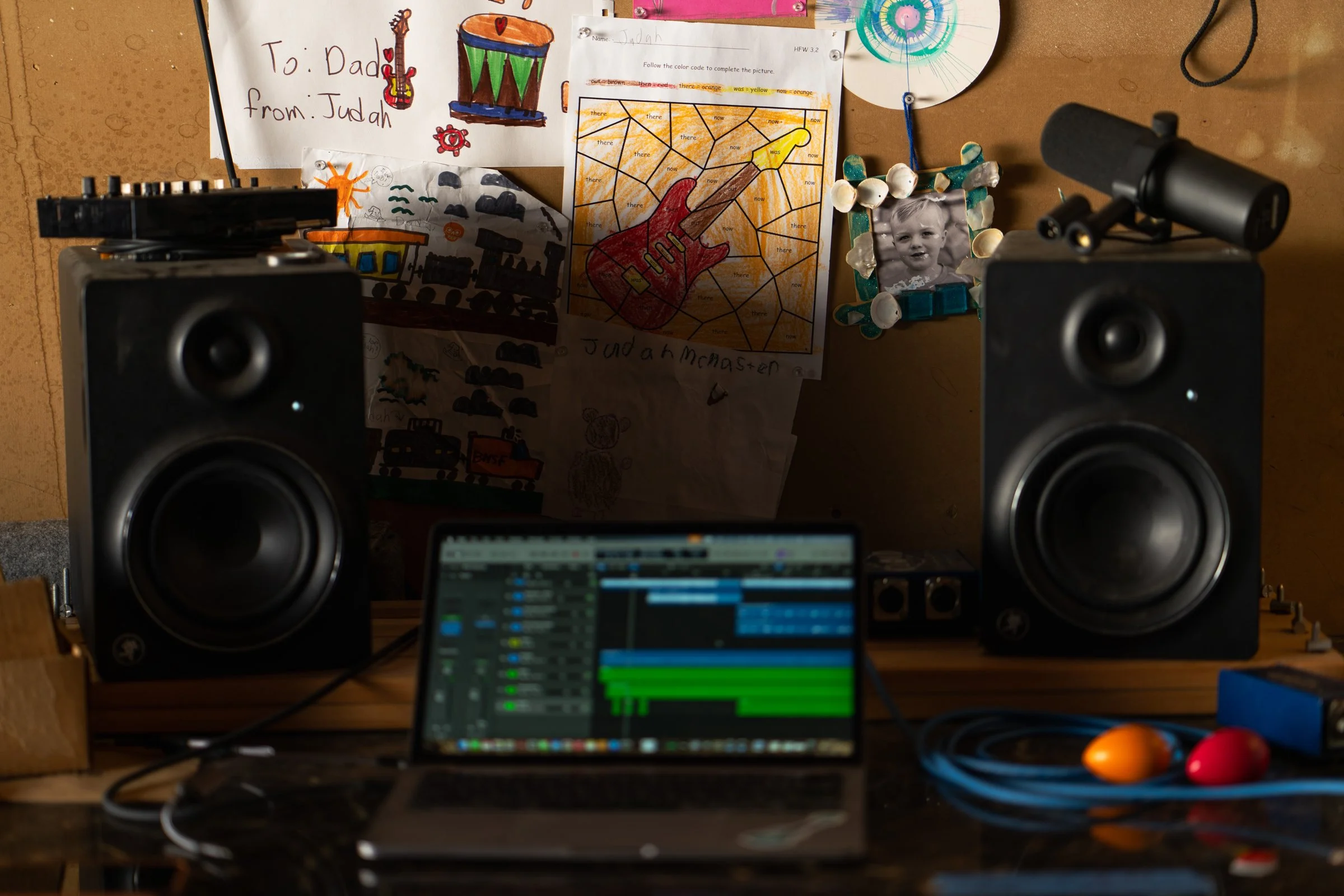 A music production setup with two large studio monitors, a laptop displaying a digital audio workstation, and children's drawings and photographs on the wall behind.