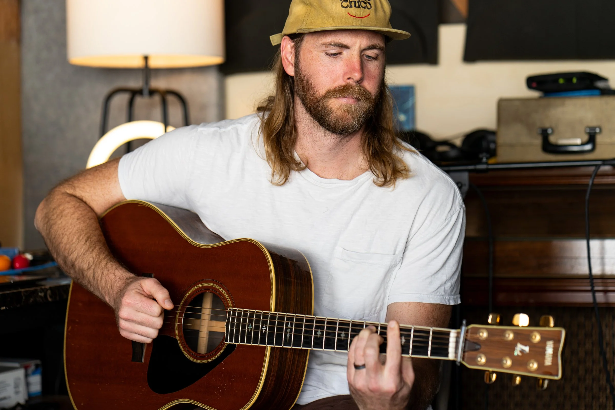 Man with long hair and beard playing acoustic guitar in a room with a lamp and equipment in the background.