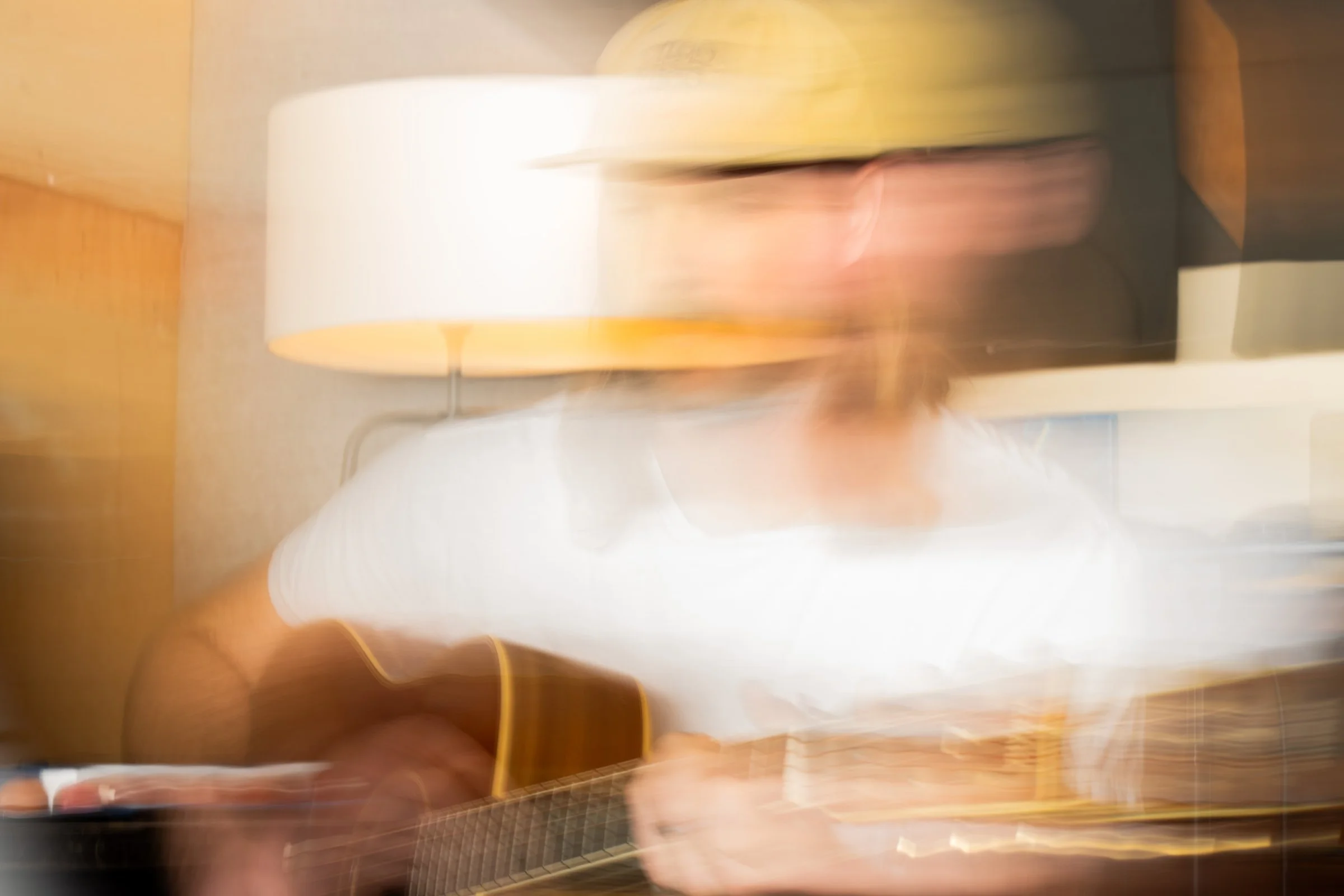Person playing an acoustic guitar in a room with a lamp and a television in the background, motion blur.