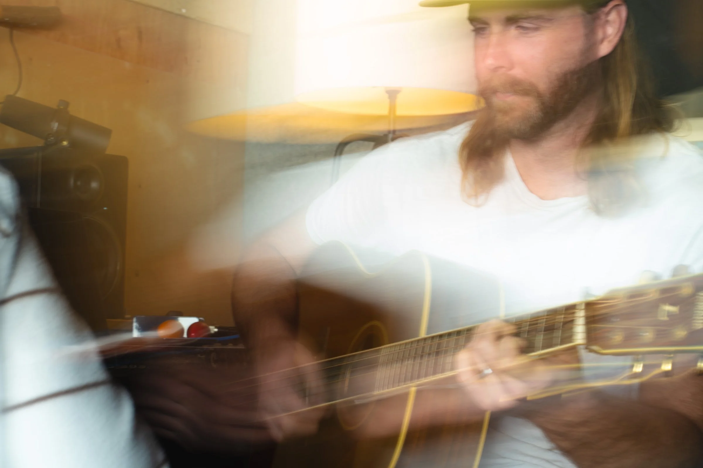 A man with long hair and a beard playing an acoustic guitar in a room with a wooden wall and a lamp.